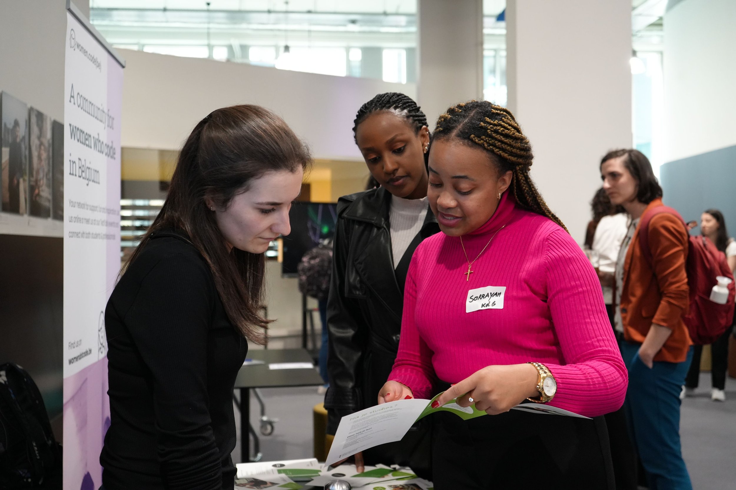 Three women are engaged in a conversation at an indoor event. One woman, wearing a bright pink top and a name tag that reads 'Soraya,' is showing a pamphlet to two other women. The background features other people and display boards.