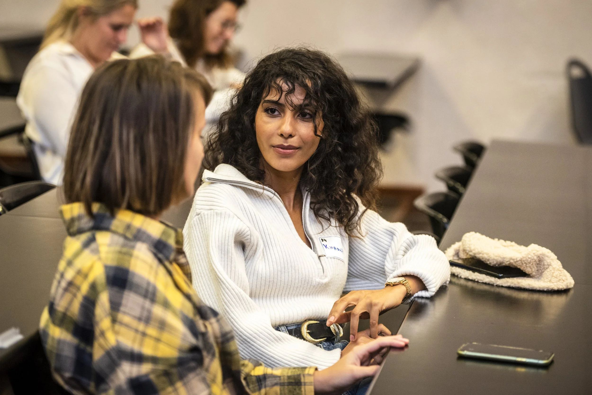 A classroom or lecture hall with students, focusing on two women sitting at a desk. One woman, with curly dark hair and a white sweater, is looking at the other woman, who has brown hair and a yellow plaid shirt, while the second woman shows something on her phone. Other students are blurred in the background.