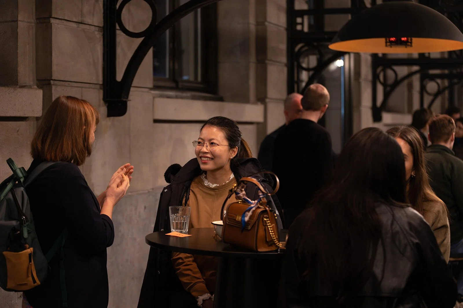 Women socializing at a bar during evening time, with people in the background and warm lighting.