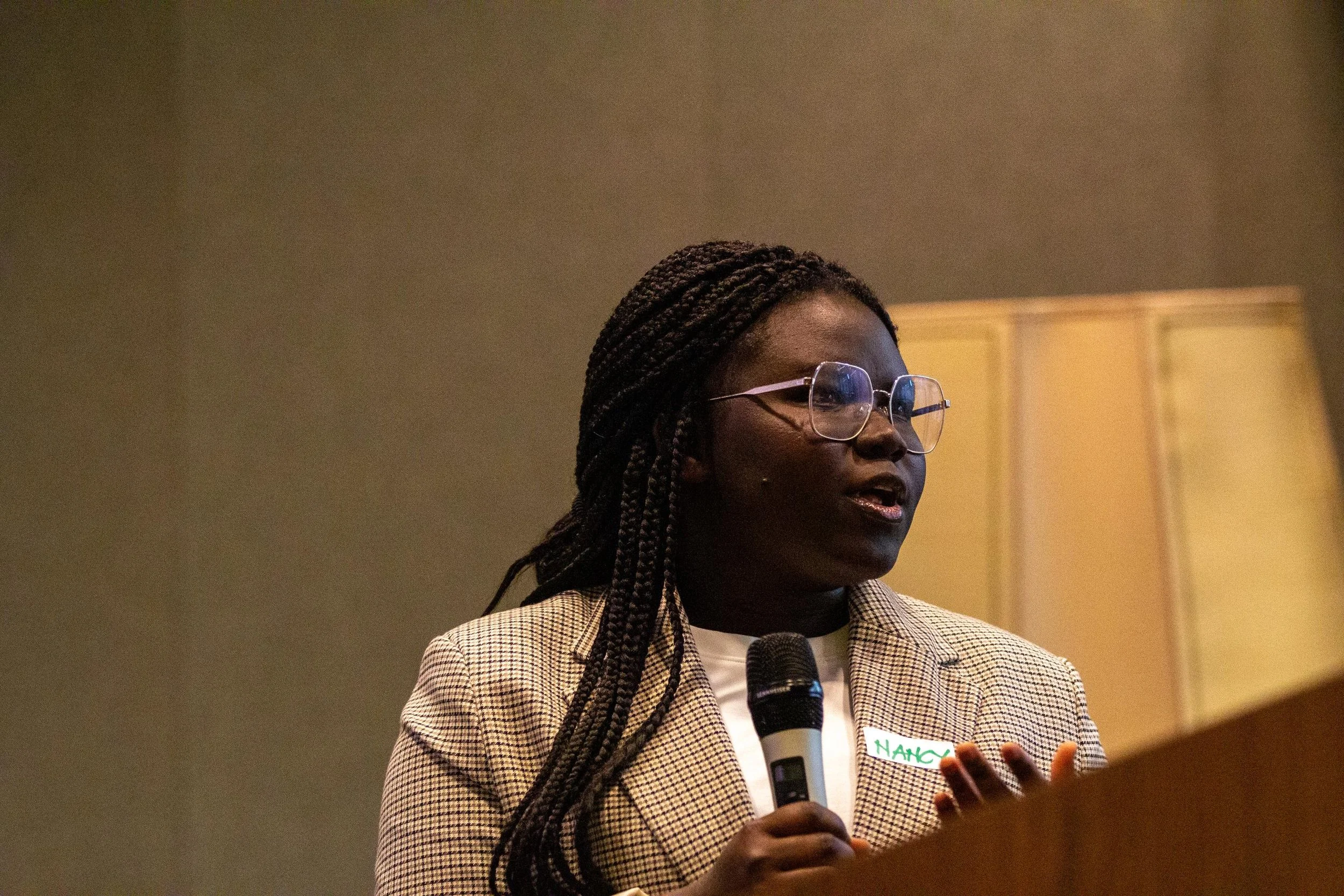 A woman with braided hair and glasses speaking into a microphone at a podium, wearing a beige blazer with a name tag.