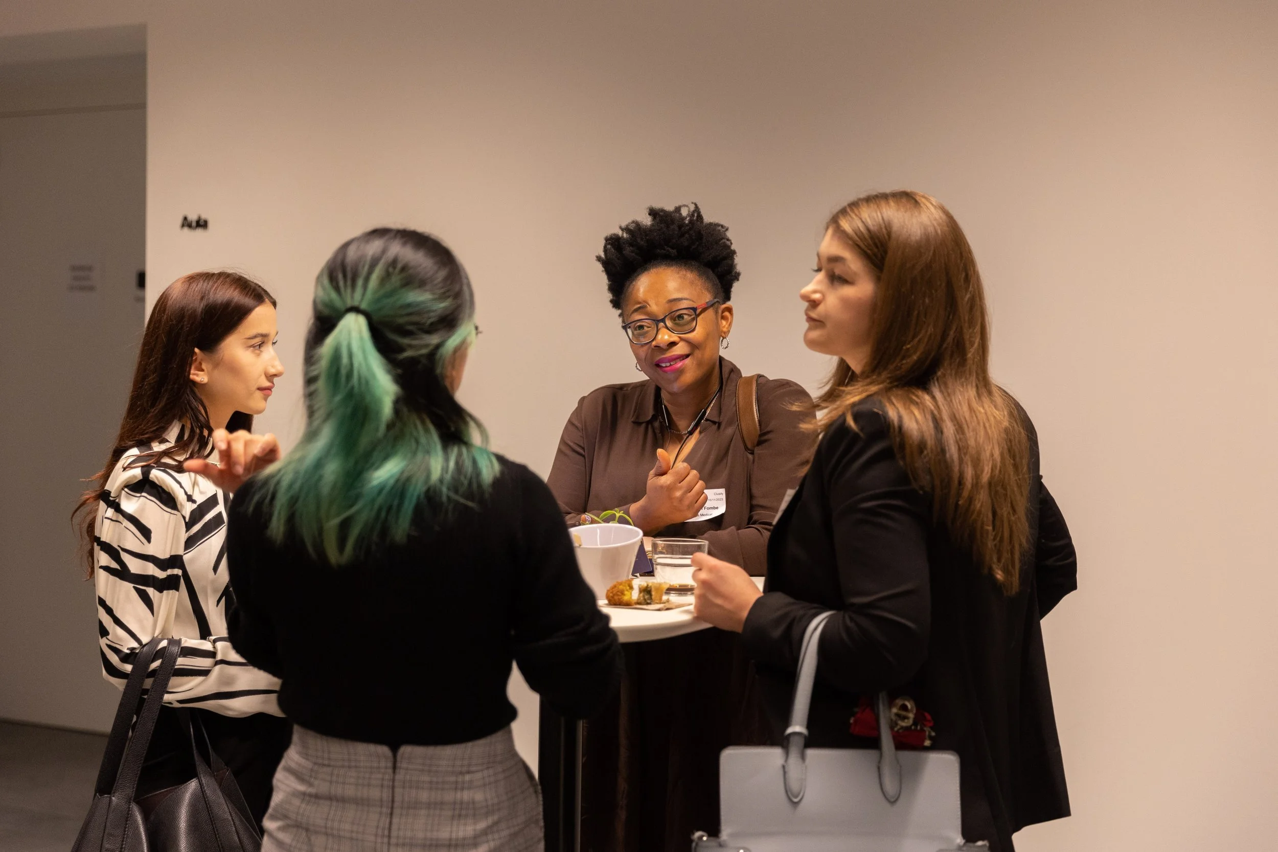 Four women standing and talking around a high table in a conference or networking event, with drinks and snacks visible on the table.