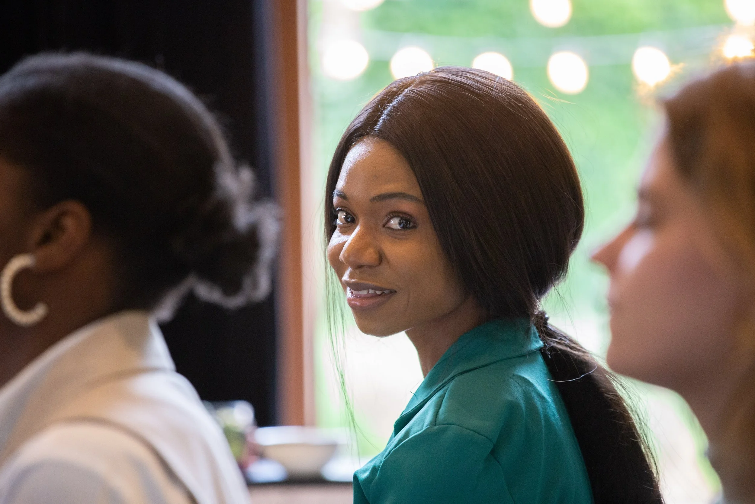 A woman with long dark hair, wearing a teal top, looking over her shoulder and smiling at the camera in a social gathering. Other women are partially visible in the foreground and background, with a blurred outside view.