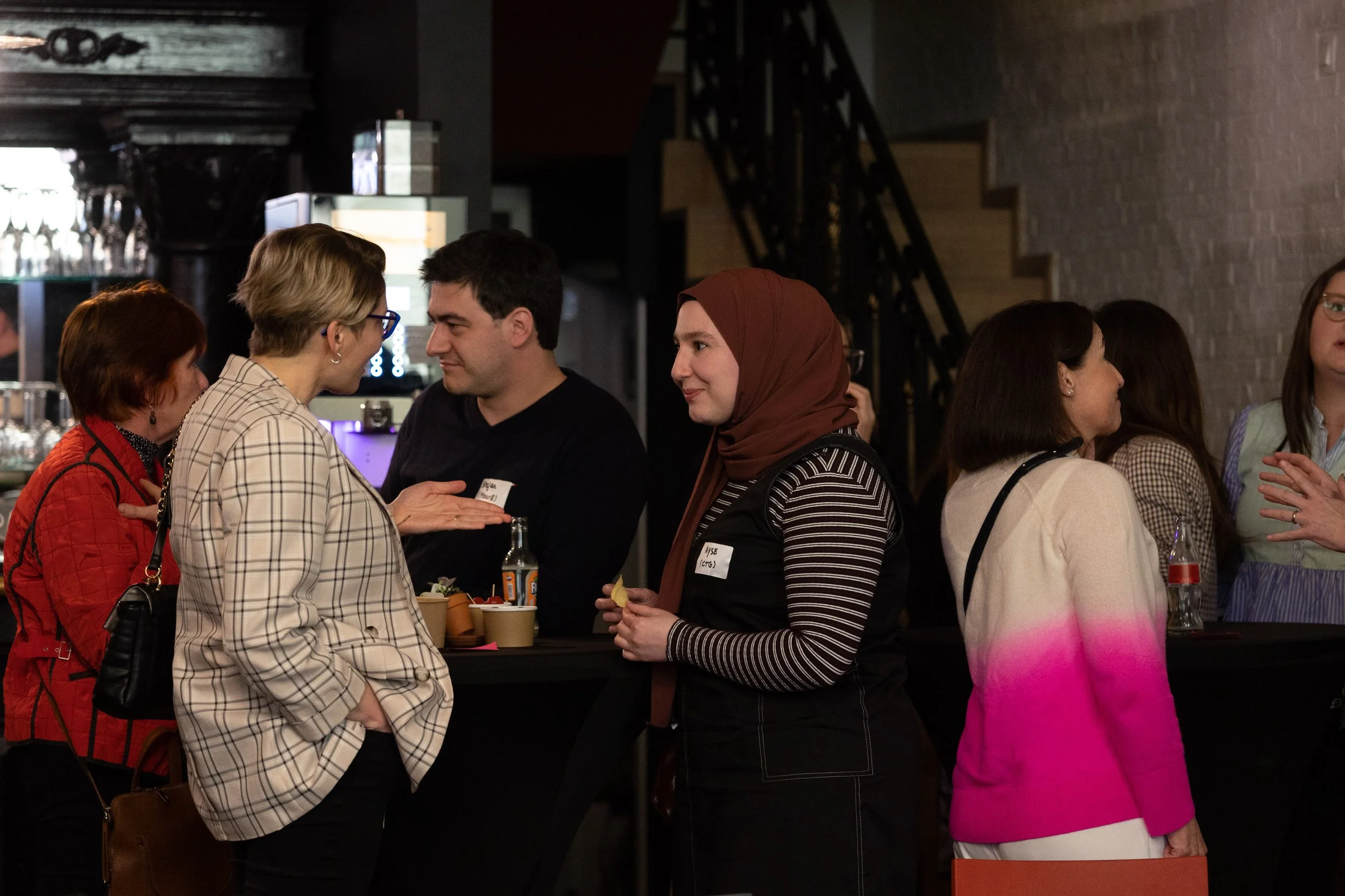 Group of people socializing at a bar or event, engaging in conversations around high tables, with drinks and snacks, in a dimly lit interior.