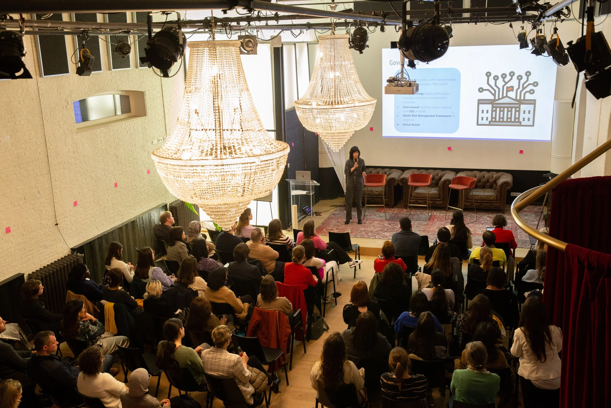 A woman giving a presentation to an audience in a conference room with large chandeliers, a stage with chairs, and a large projection screen displaying a slide with text and graphics.
