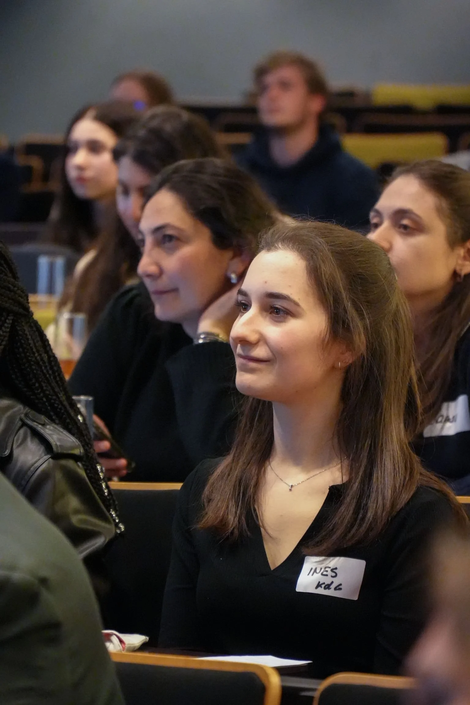 Young woman with brown hair wearing a black top and a name tag that says "INES" at a conference or seminar.