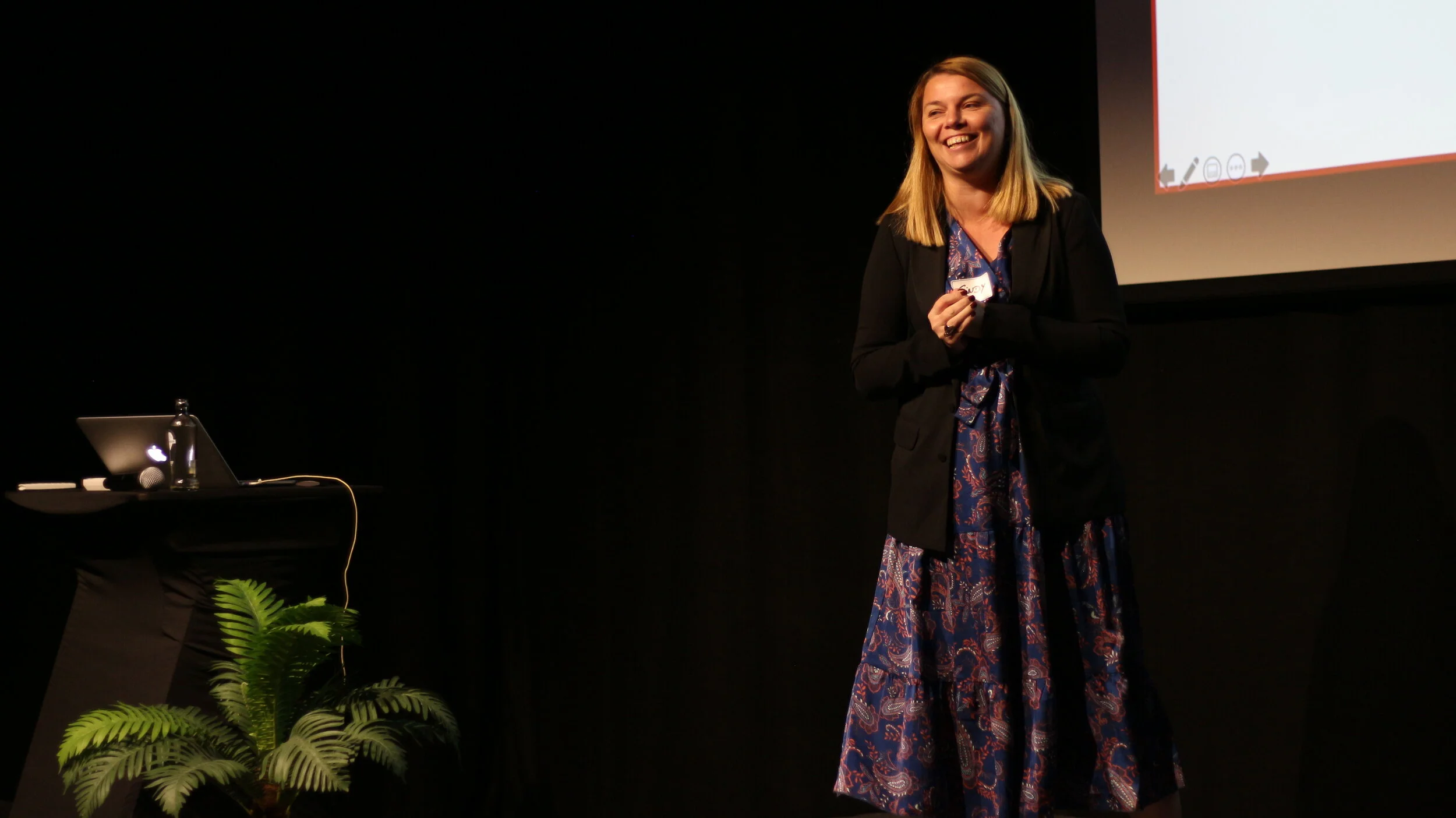 A woman standing on a stage, smiling, holding a small object, with a large presentation screen behind her, a laptop, water bottle, and microphone on a table nearby, and a potted plant in front of the table.