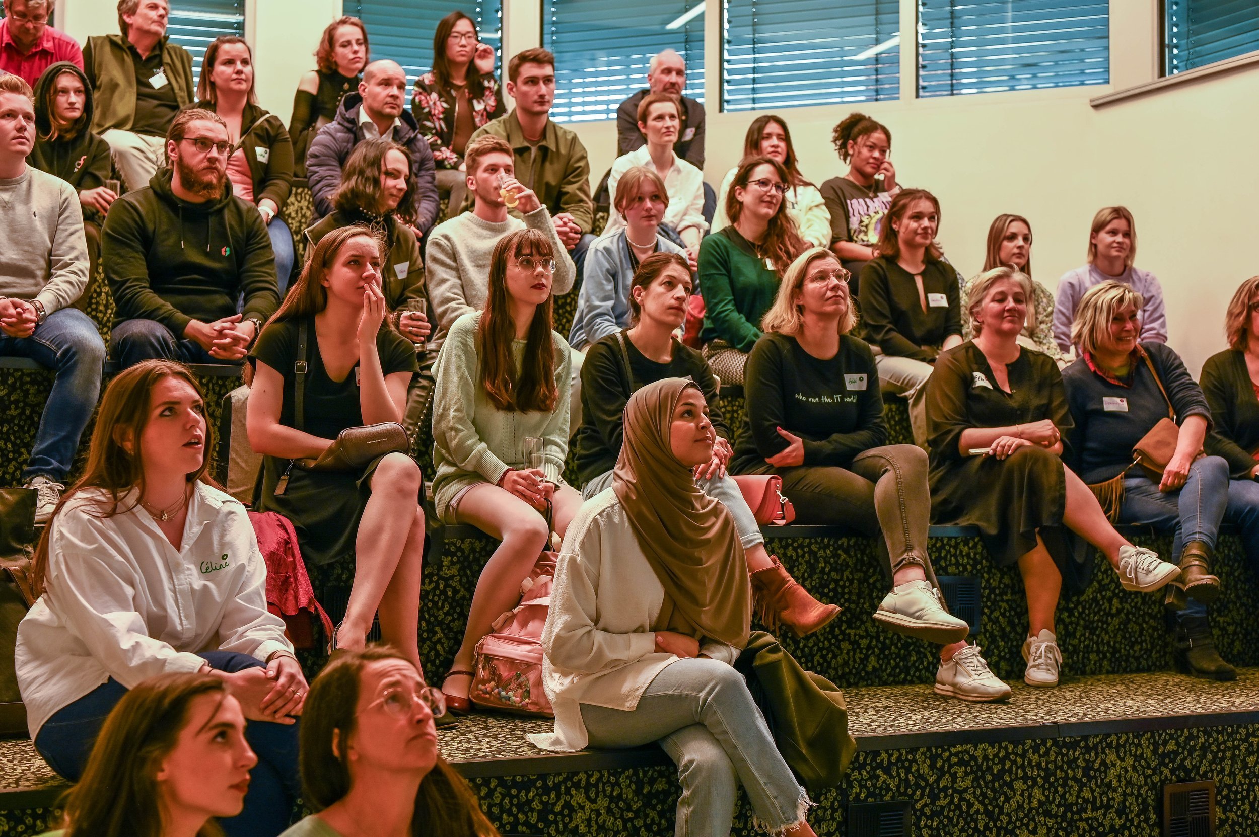 A group of diverse people seated on tiered black and yellow patterned seating, attentively watching a presentation or performance in a room with green walls and blue window blinds.