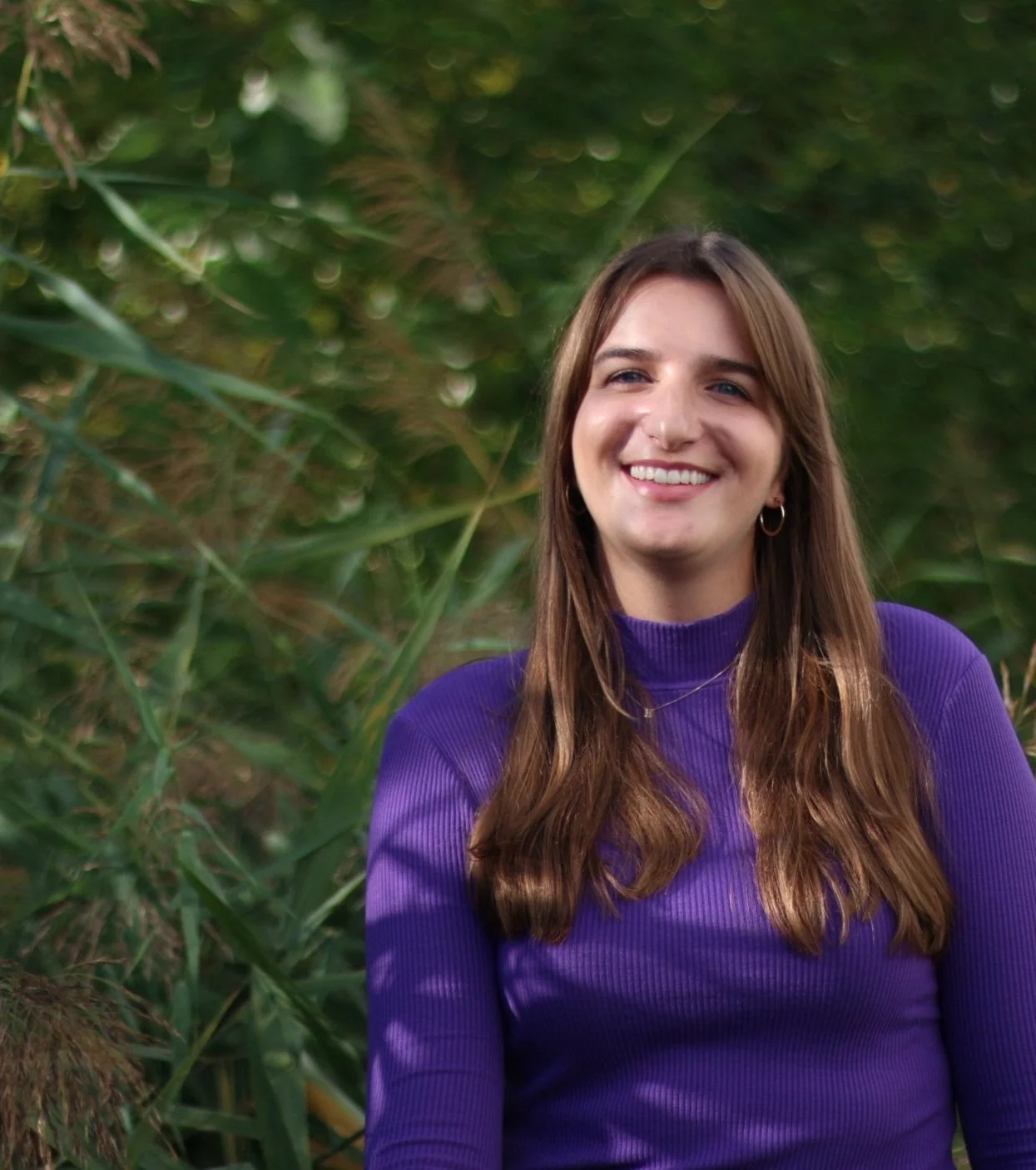 A young woman with long brown hair wearing a purple long-sleeve top smiling outdoors in a green, leafy environment.