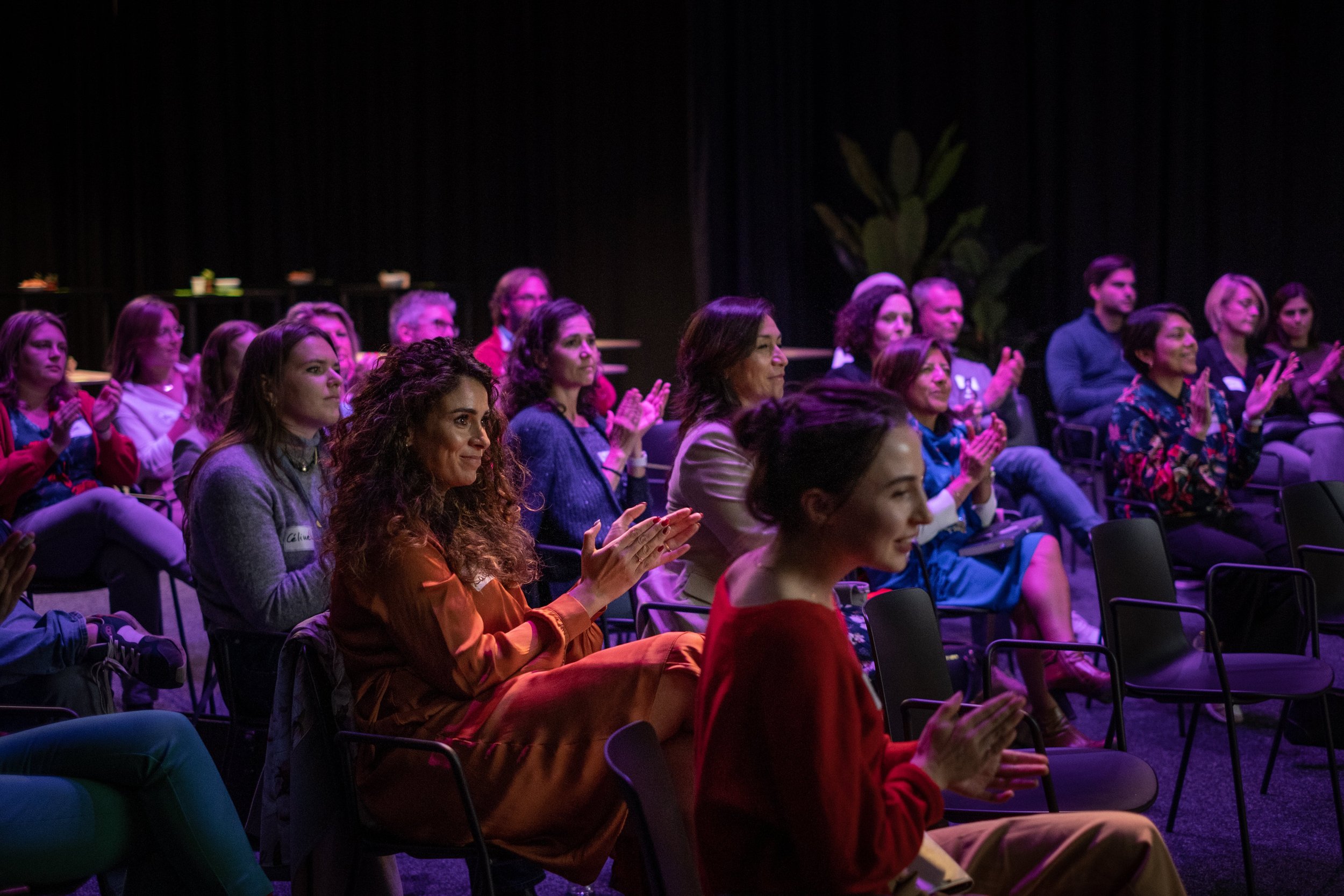 Audience members seated and clapping at an indoor event with a dark background and some greenery.