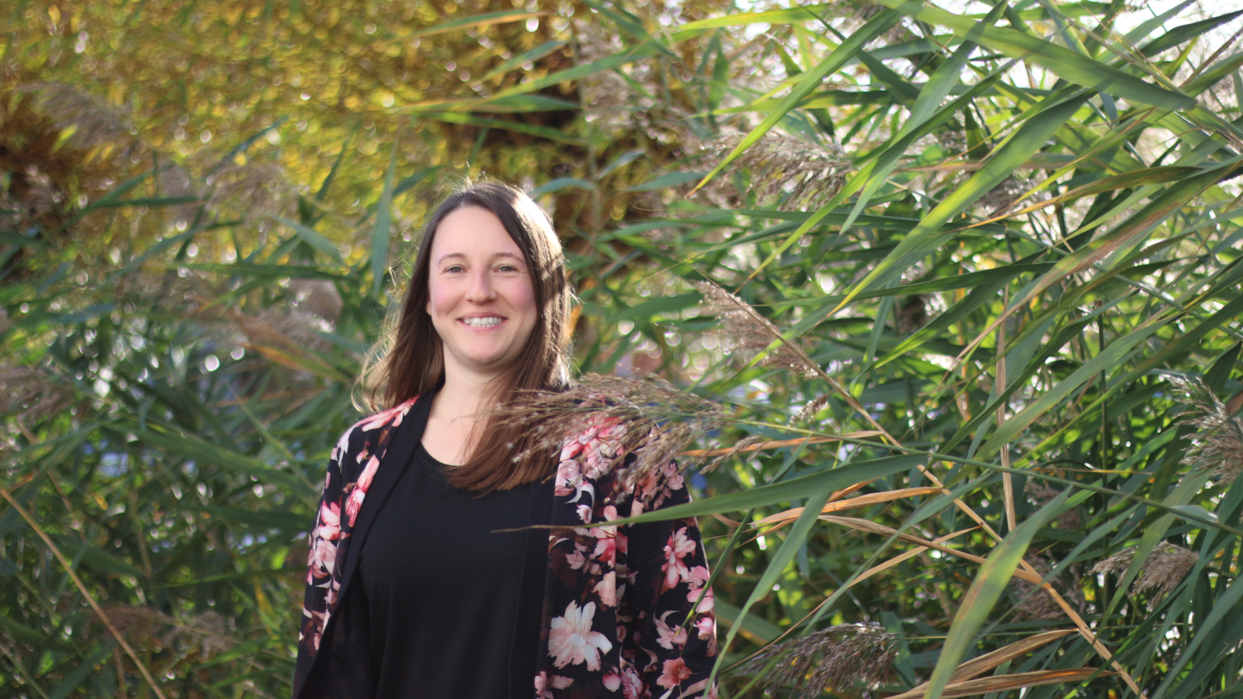 A woman smiling and standing in a lush, green outdoor setting surrounded by tall grasses and plants.
