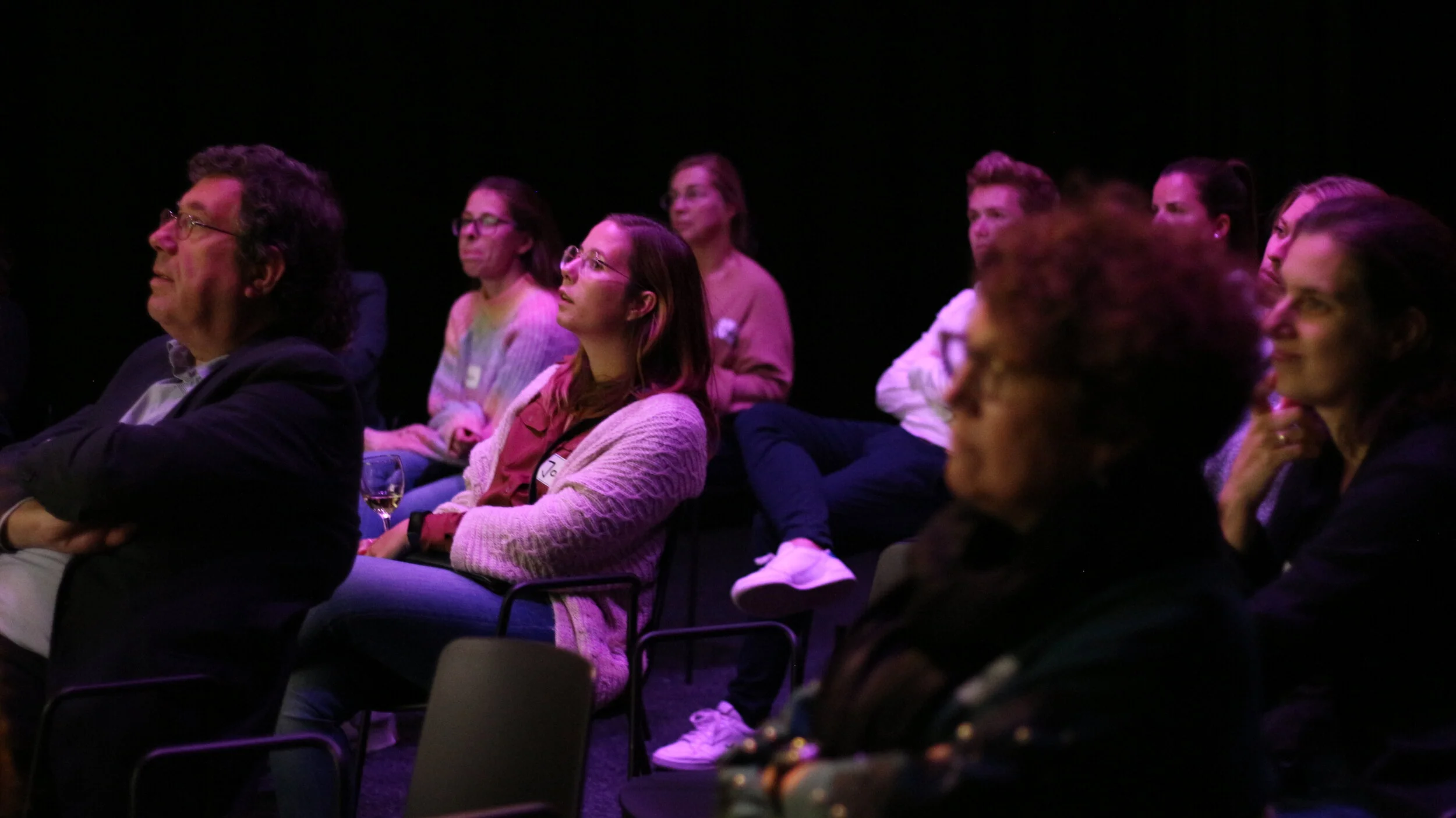Audience of diverse adults seated and attentively listening at an indoor event or presentation.