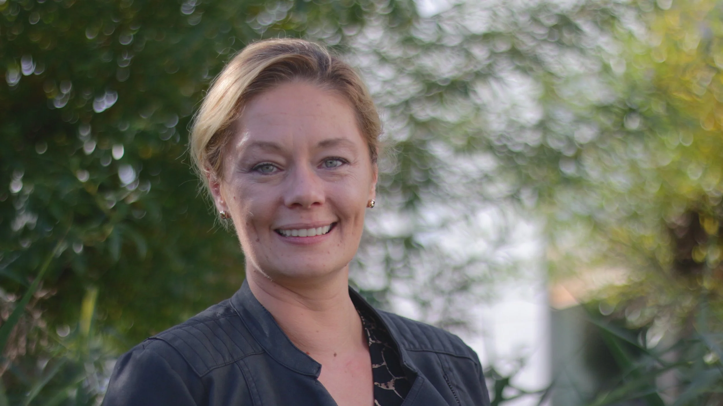A woman with short blonde hair and blue eyes smiling outdoors with greenery in the background.
