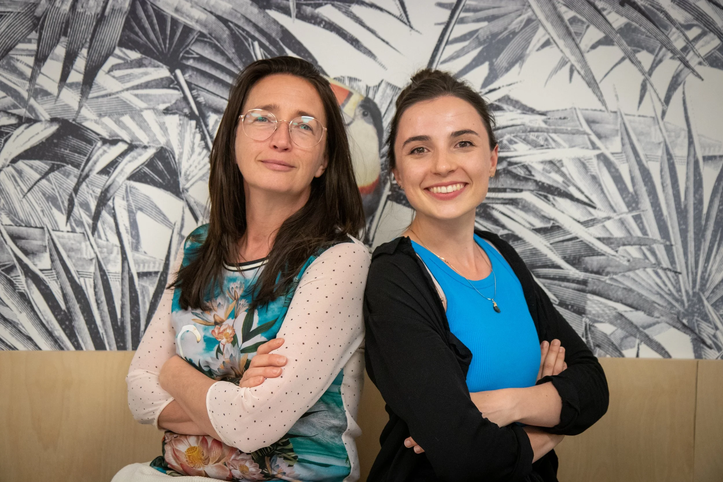 Two women standing back to back with crossed arms, smiling, in front of a black and white mural of tropical leaves and a toucan.