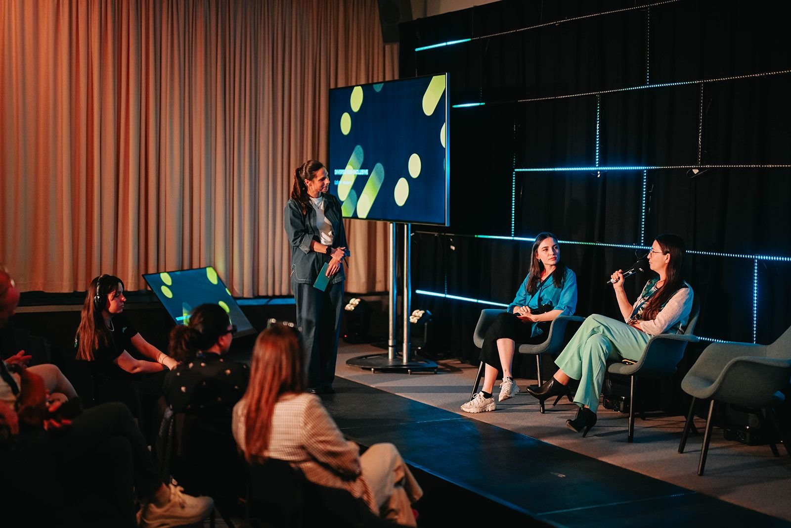 Women participating in a panel discussion at a conference with an audience watching. The stage has large screens and modern lighting.
