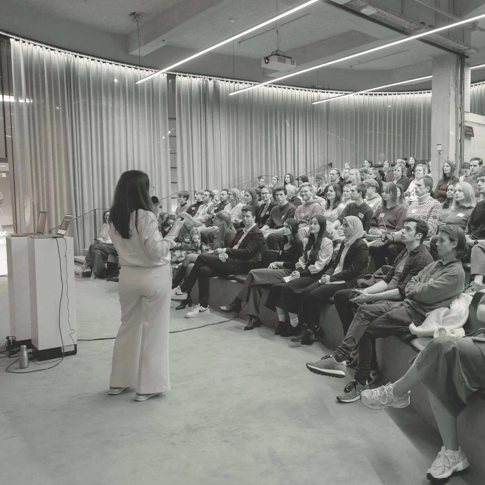 A woman giving a presentation to an attentive audience in a modern conference room with curtains and overhead lighting.