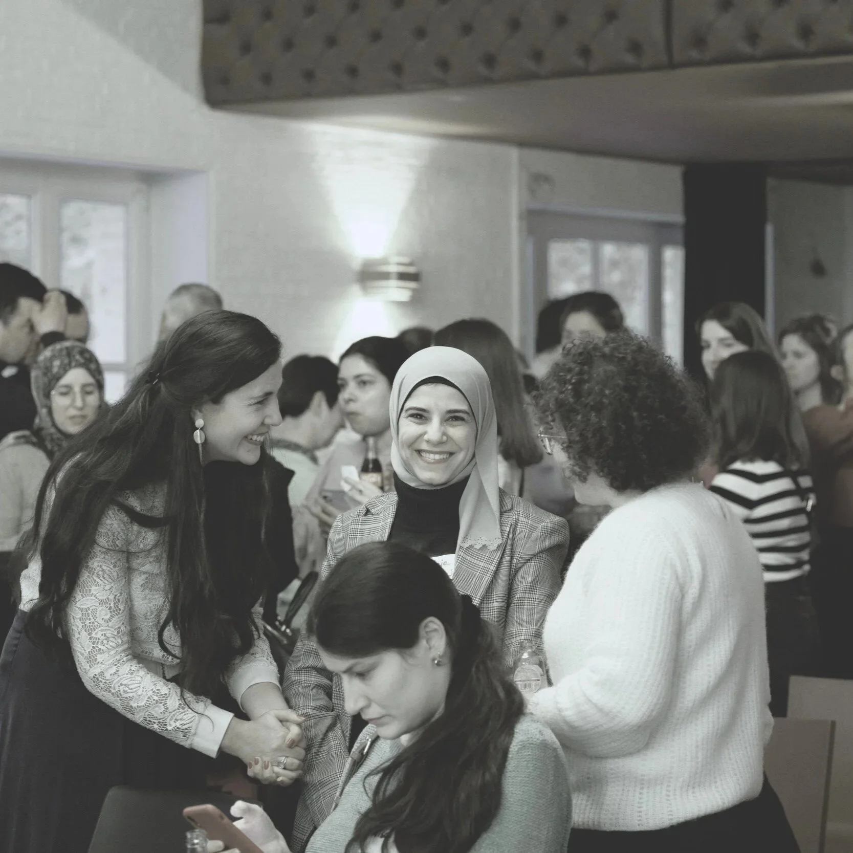 A group of diverse women socializing at an indoor event, including a smiling woman wearing a hijab, with some women chatting and others looking at their phones.