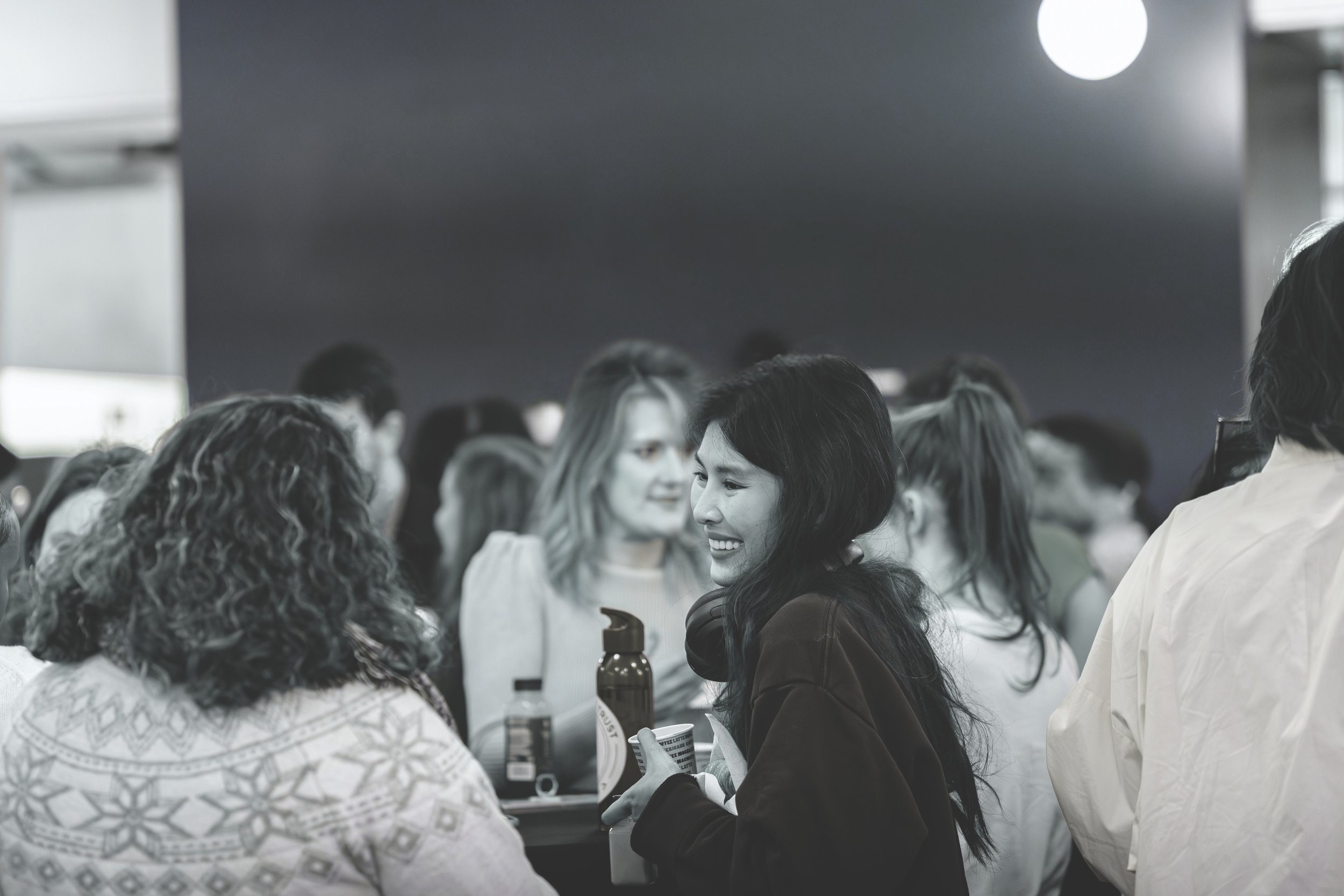 A group of people at a social gathering, with a woman with long dark hair smiling and holding a bottle, surrounded by others in conversation.