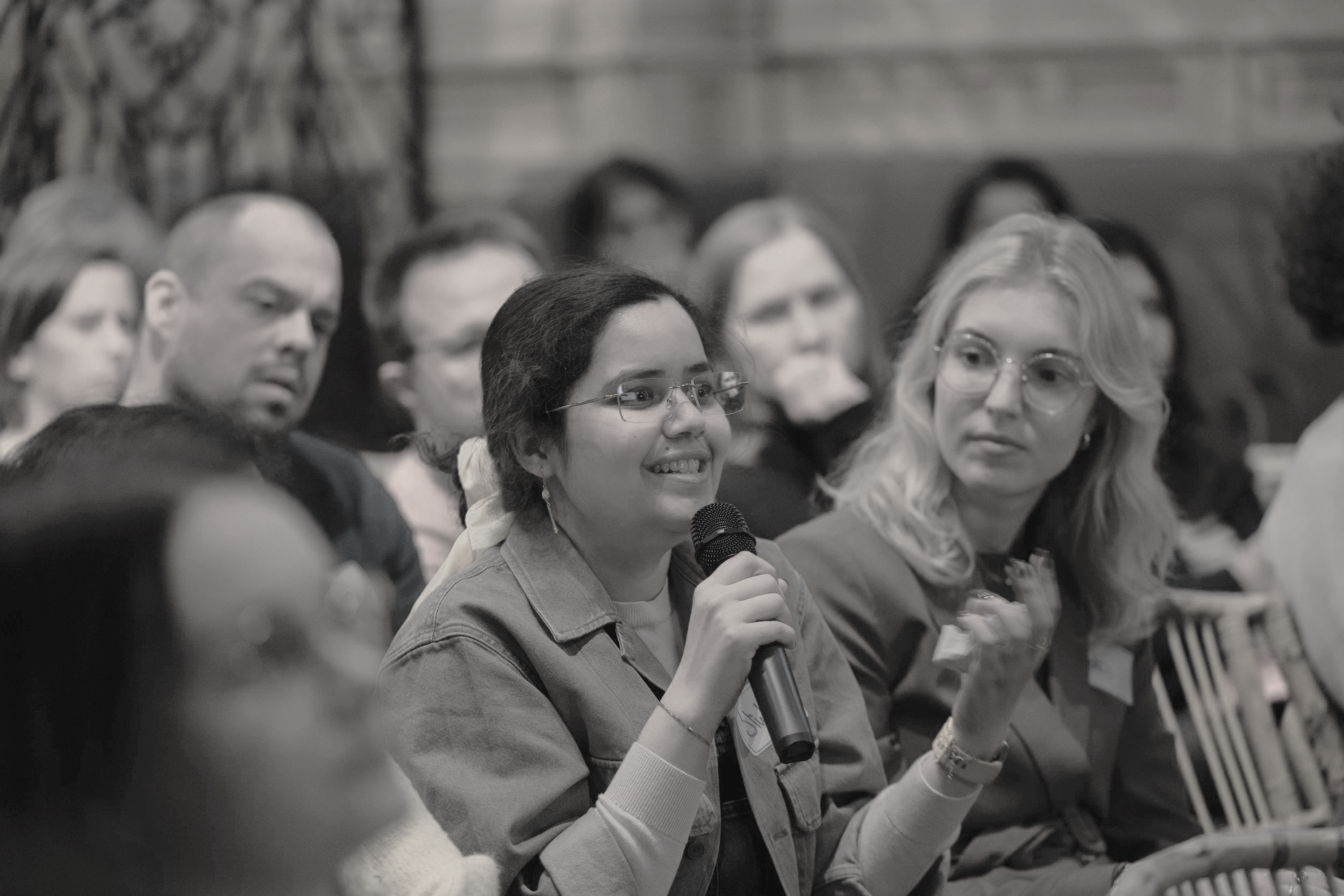 A woman with glasses speaking into a microphone at a conference or meeting, with an attentive audience around her.