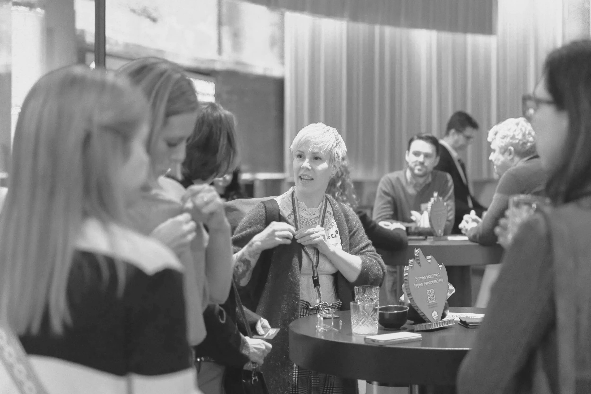 A group of people at an indoor event, with a woman with short hair engaging in conversation. Others are sitting or standing around tables, some with drinks and papers, in a well-lit setting.