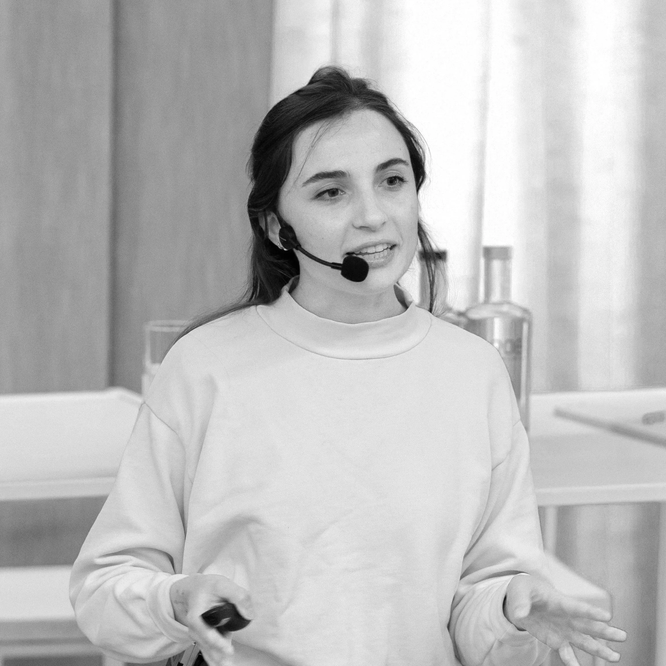 A young woman wearing a headset and microphone, gesturing with her hands, giving a presentation or speech.