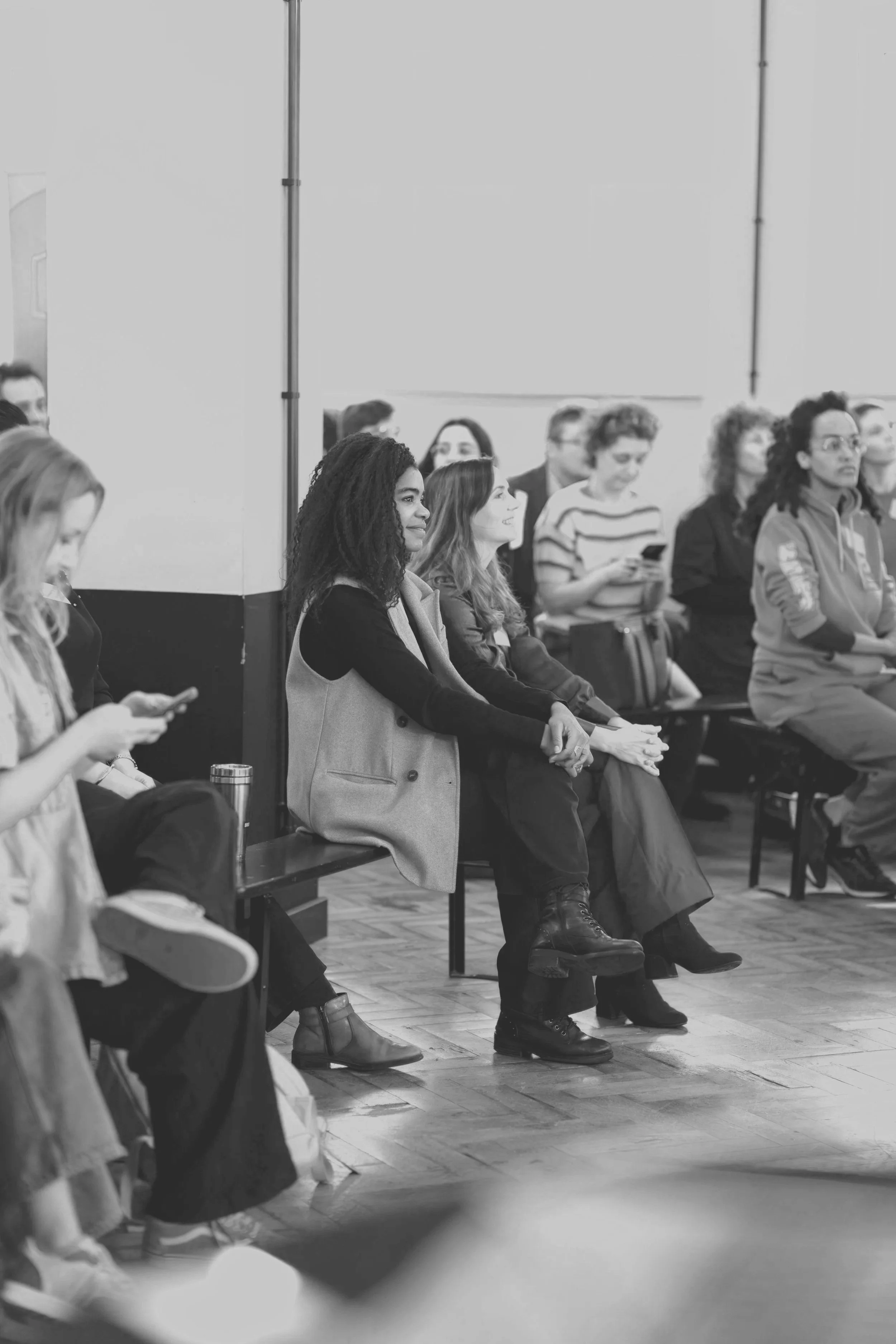 A group of diverse people sitting on benches in an indoor setting, some using mobile phones, during a formal event.