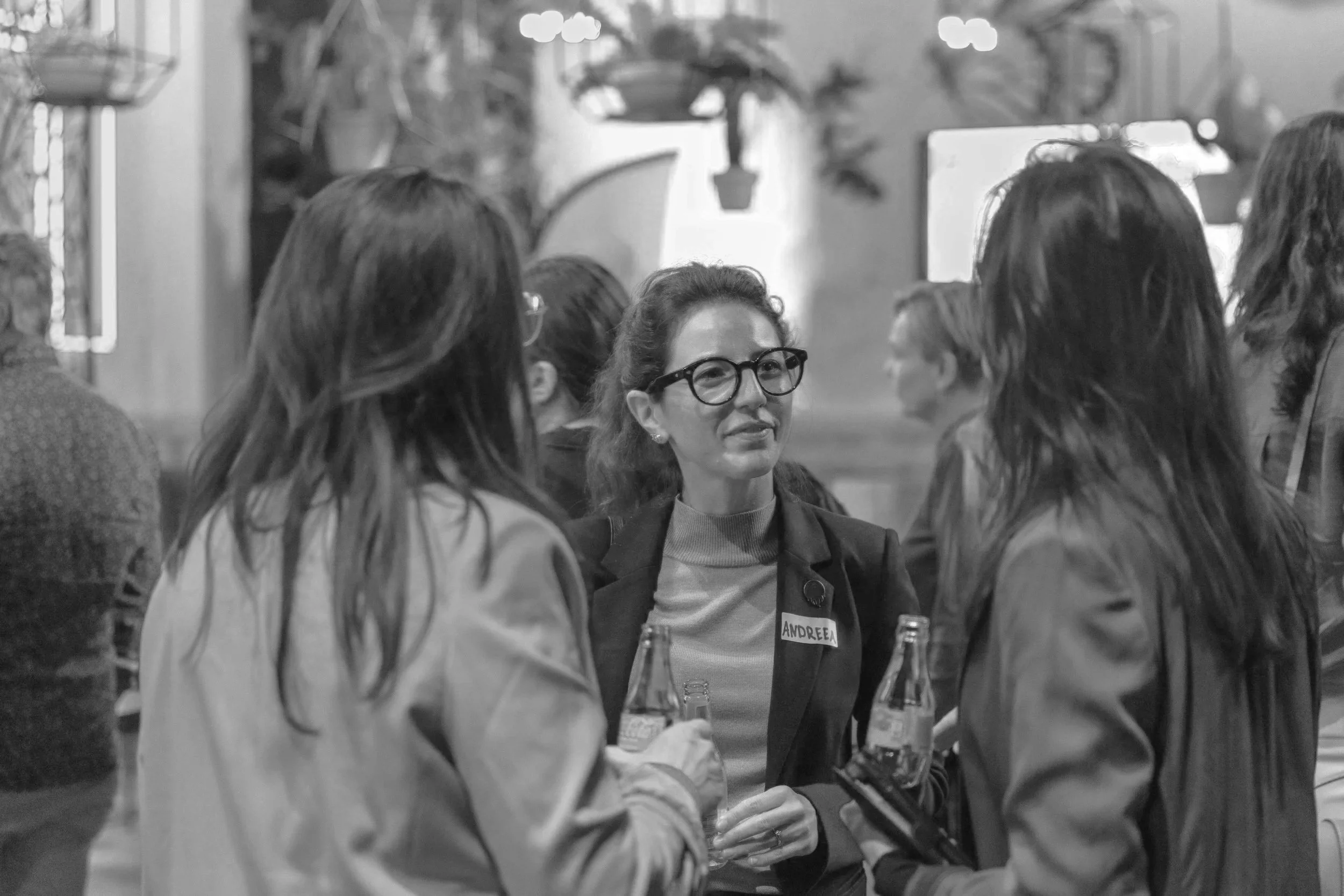 Group of women socializing indoors, one woman wearing glasses and a name tag that says 'ANDREA' in the center.