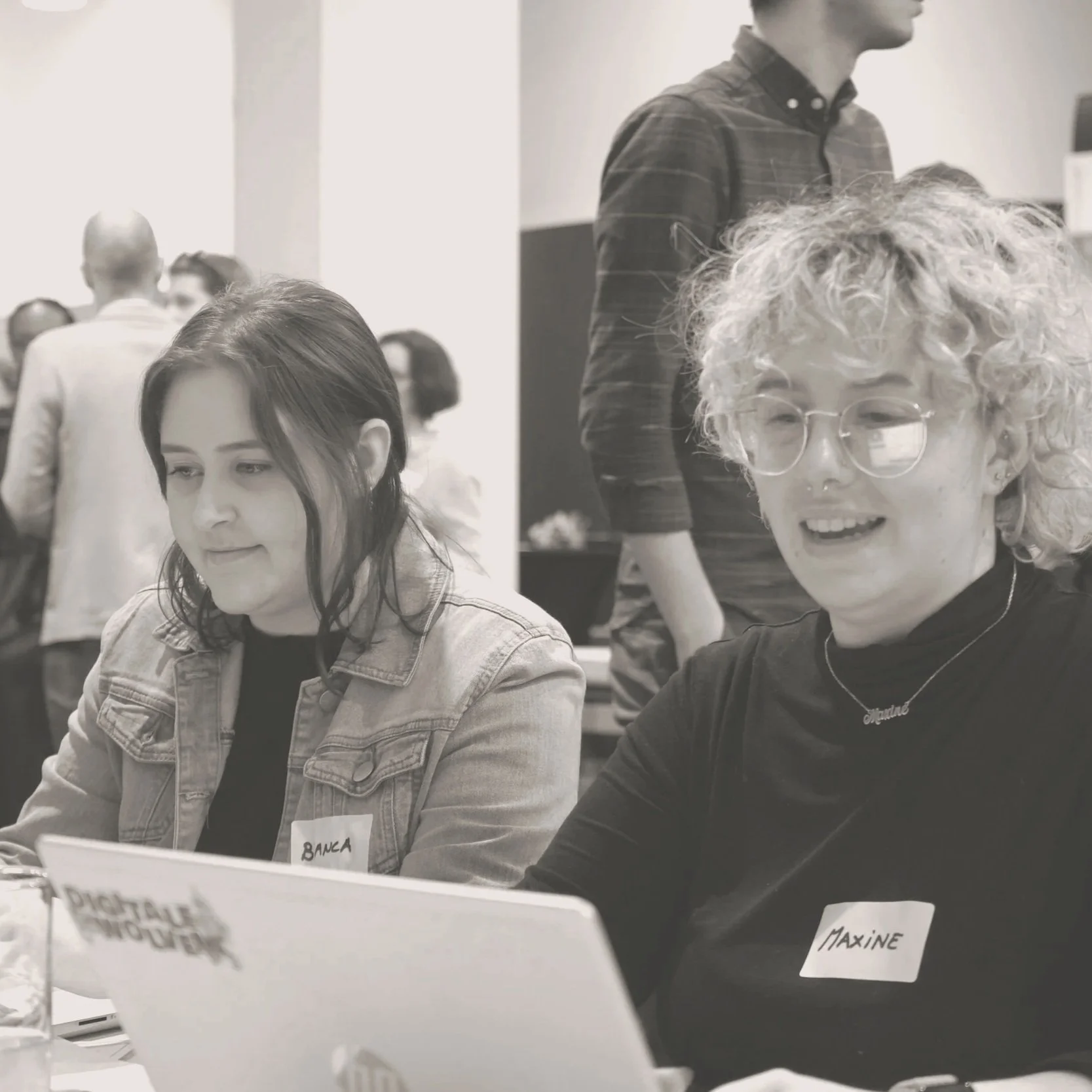 Two women sitting at a table, working on laptops, in a busy environment. One woman has dark hair and a denim jacket, with a name tag that says 'BANA.' The other woman has curly blonde hair, glasses, and a necklace that says 'Stephanie,' with a name tag that says 'Maxine.'