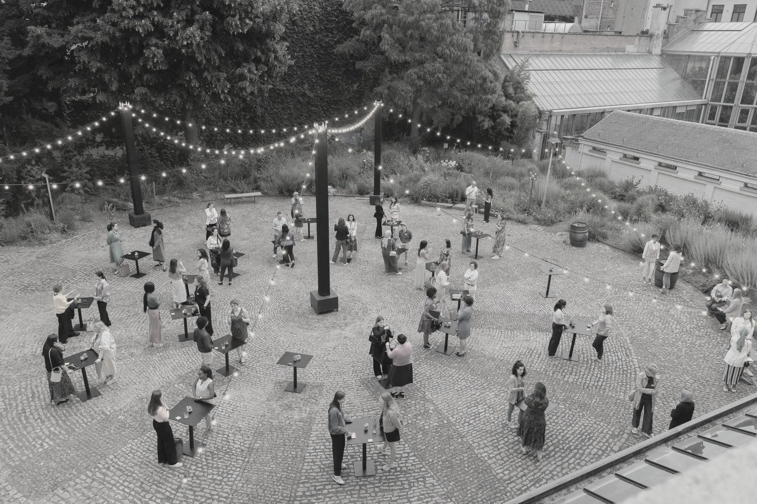Outdoor social gathering with multiple small groups of people standing at high-top tables, socializing, under string lights at night in a courtyard with cobblestone paving and surrounding greenery.