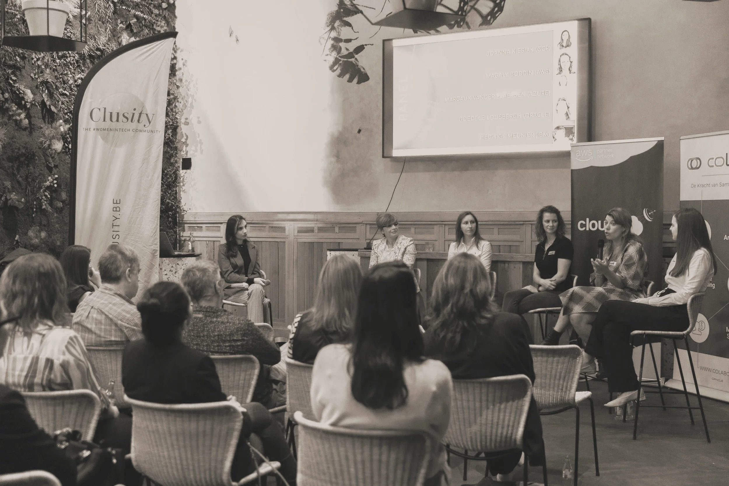 Women panel discussion at a conference, with an audience seated in front. Banners and a large screen are visible in the background.