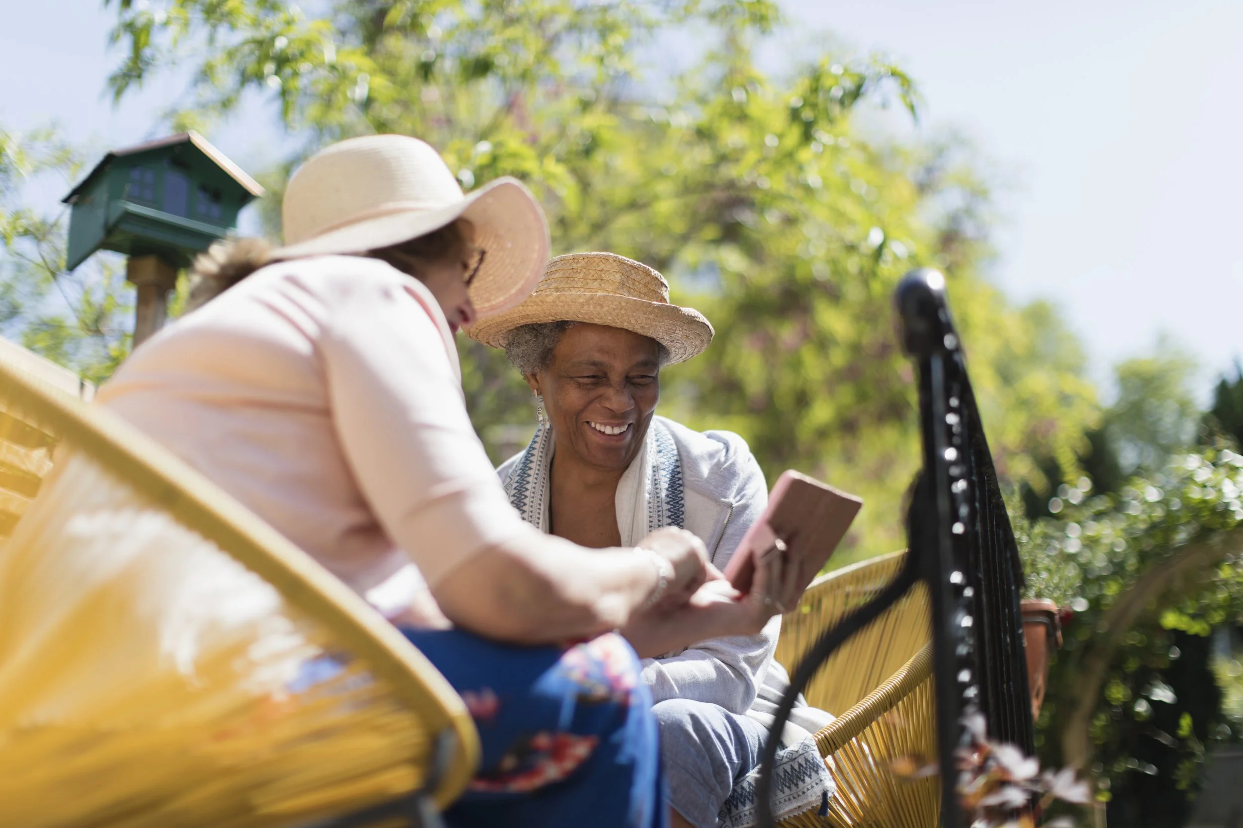 Two women sitting outside on a yellow bench, wearing sun hats and clothing, looking at a smartphone, with greenery and a birdhouse in the background on a sunny day.