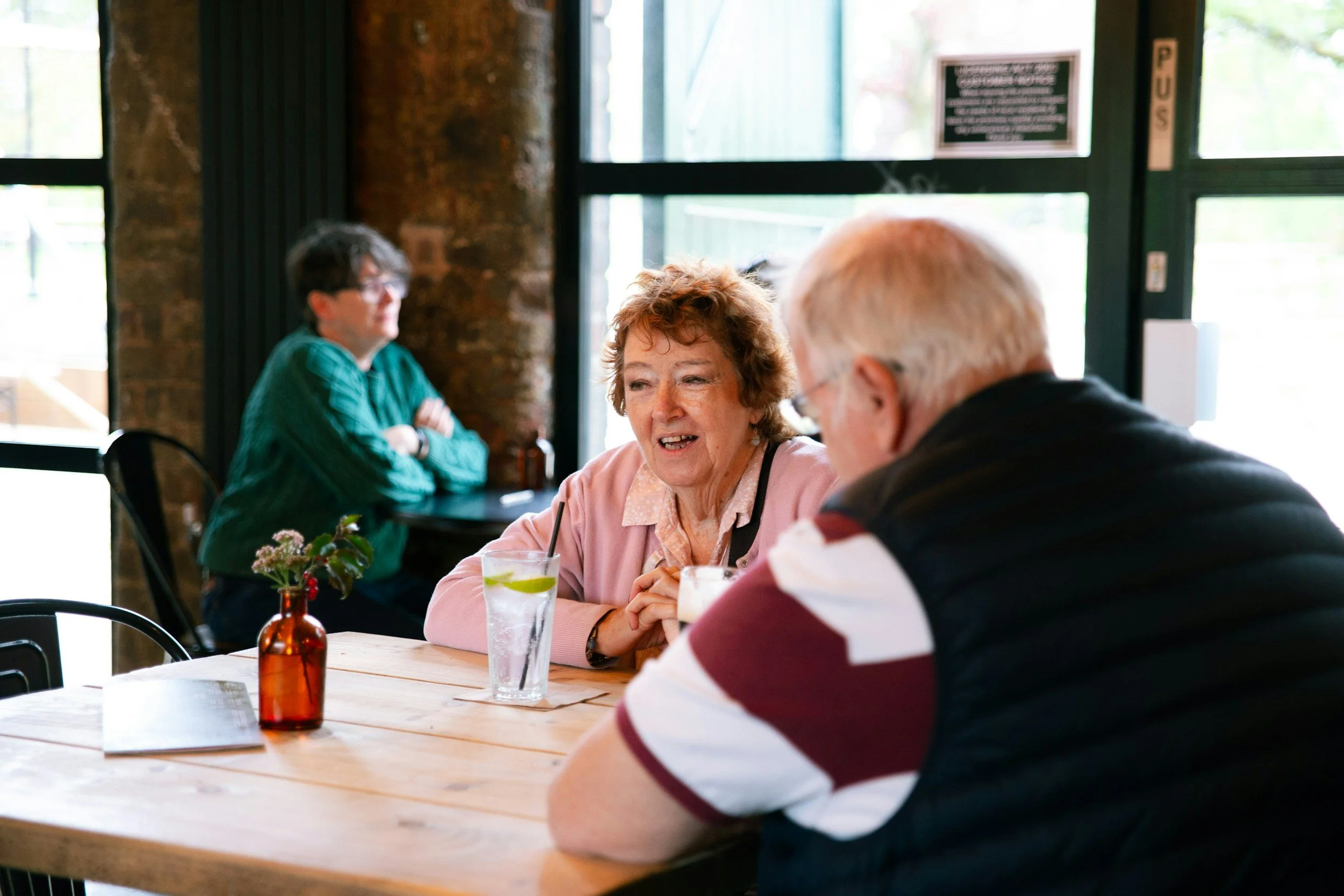 Three people sitting at a wooden table in a cafe, two women and one man, engaged in conversation, with drinks and a small flower vase on the table.