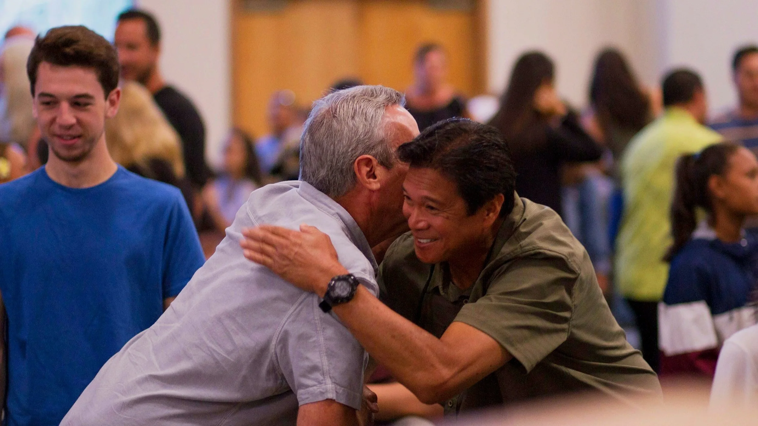 Two men greet each other with a hug at a crowded indoor event, smiling warmly, with other people in the background.