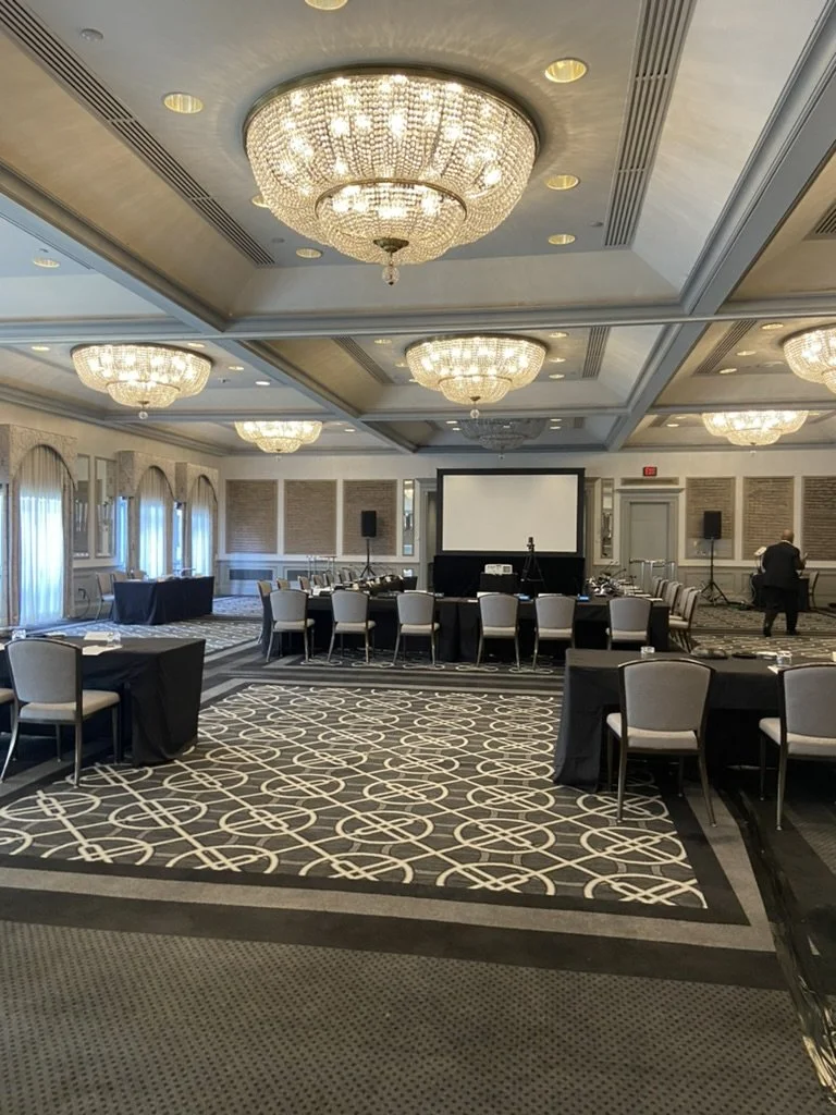 A large, elegant conference room with multiple round tables covered with black tablecloths, surrounded by chairs, and a central presentation area with a large screen and sound equipment. The ceiling features grand chandeliers, and the room has decorated walls with large windows draped with curtains.