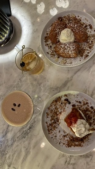 Two plates with cinnamon roll desserts topped with powdered sugar and whipped cream, a glass of hot chocolate with whipped cream, a glass of iced tea with lemon, and a cup of coffee on a marble table.
