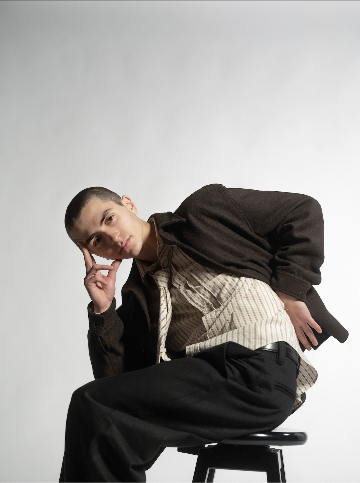 A young man sitting on a stool, wearing a black jacket, a striped shirt, and black pants, leaning forward with his right hand resting on his face and his left hand on his back, against a plain white background.