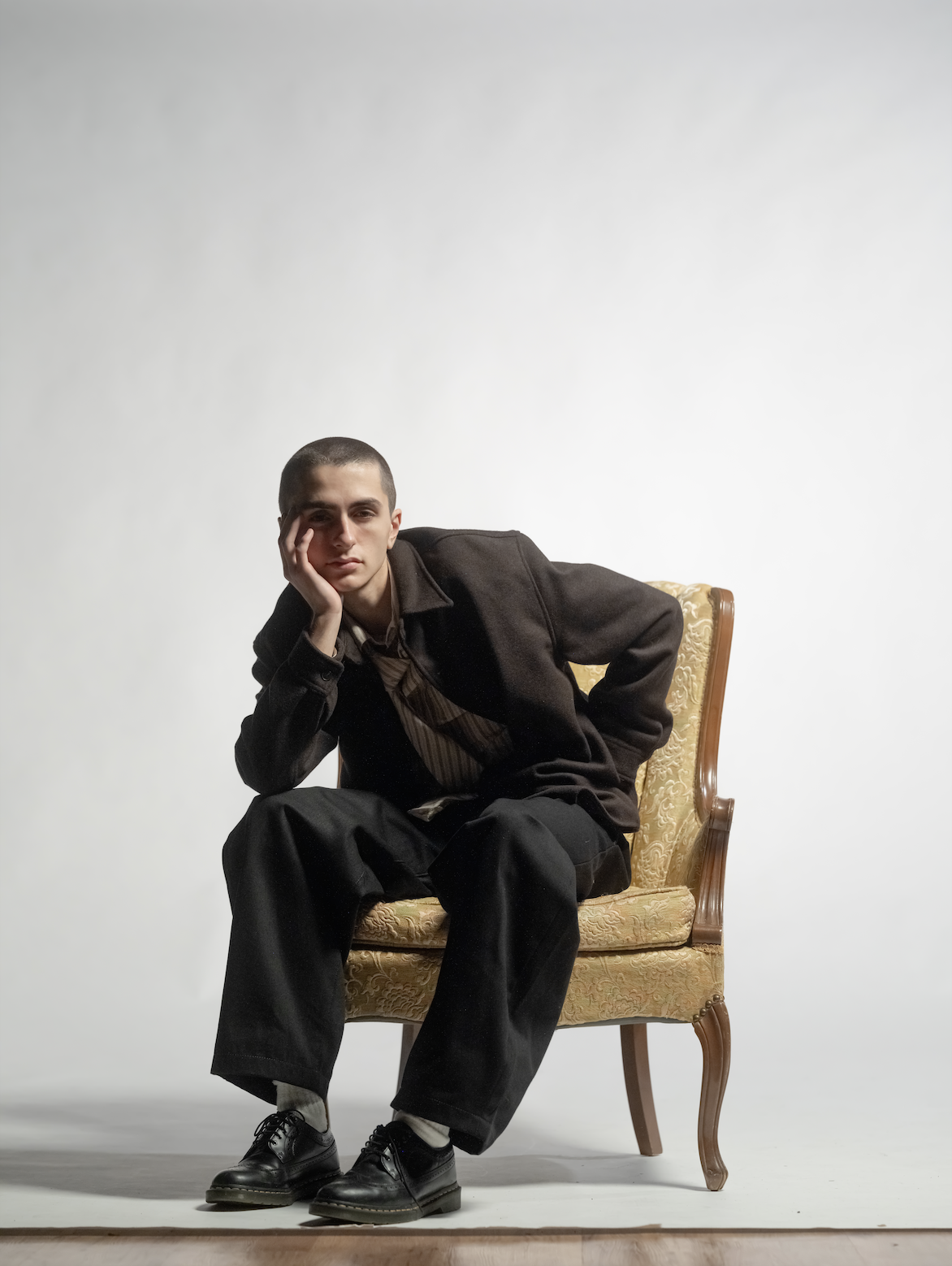 Young man sitting on a vintage upholstered armchair with one hand resting on his face against a plain white background.