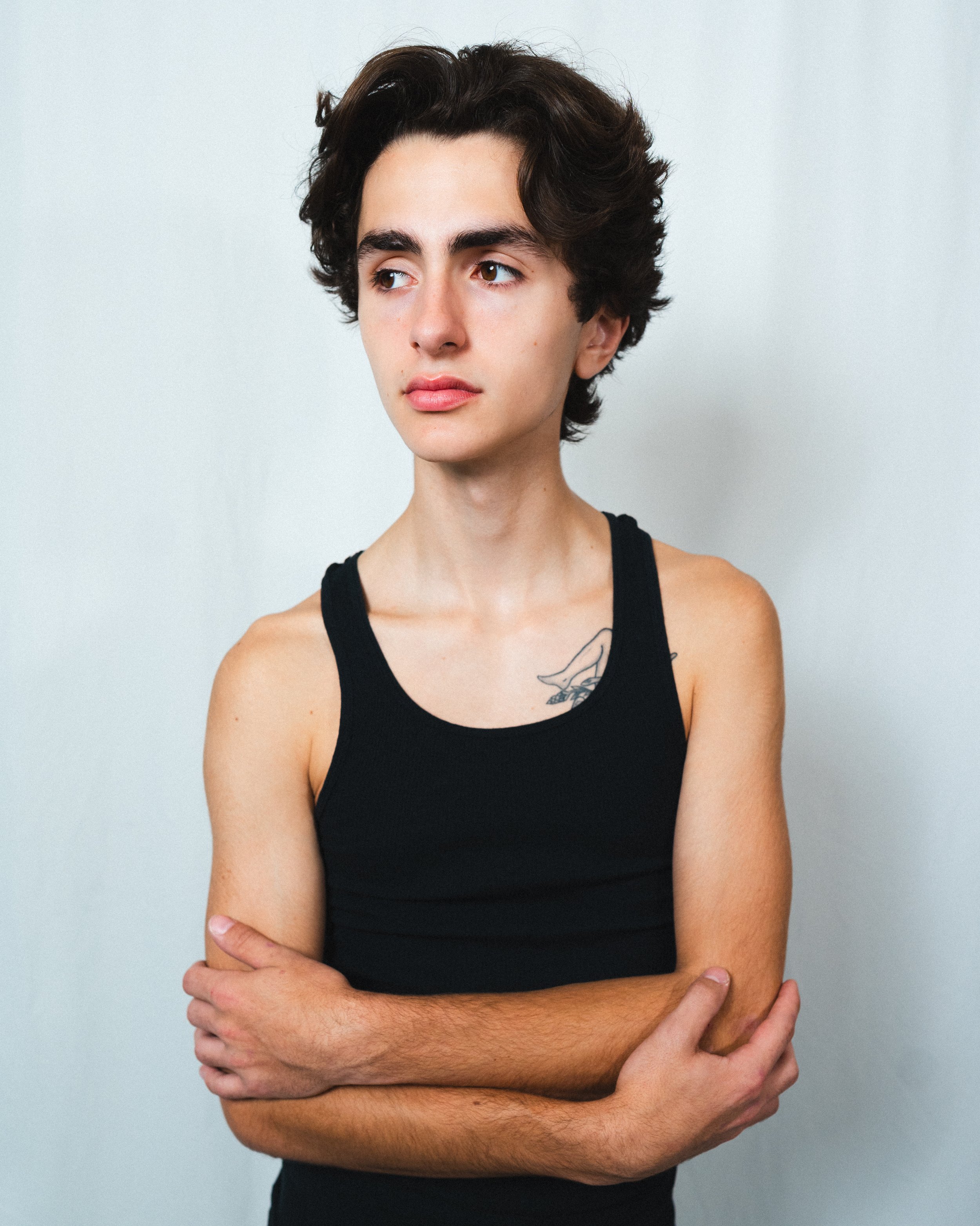 Young man with short dark hair wearing a black tank top, standing against a light background, looking to the side, with a tattoo on his left shoulder.