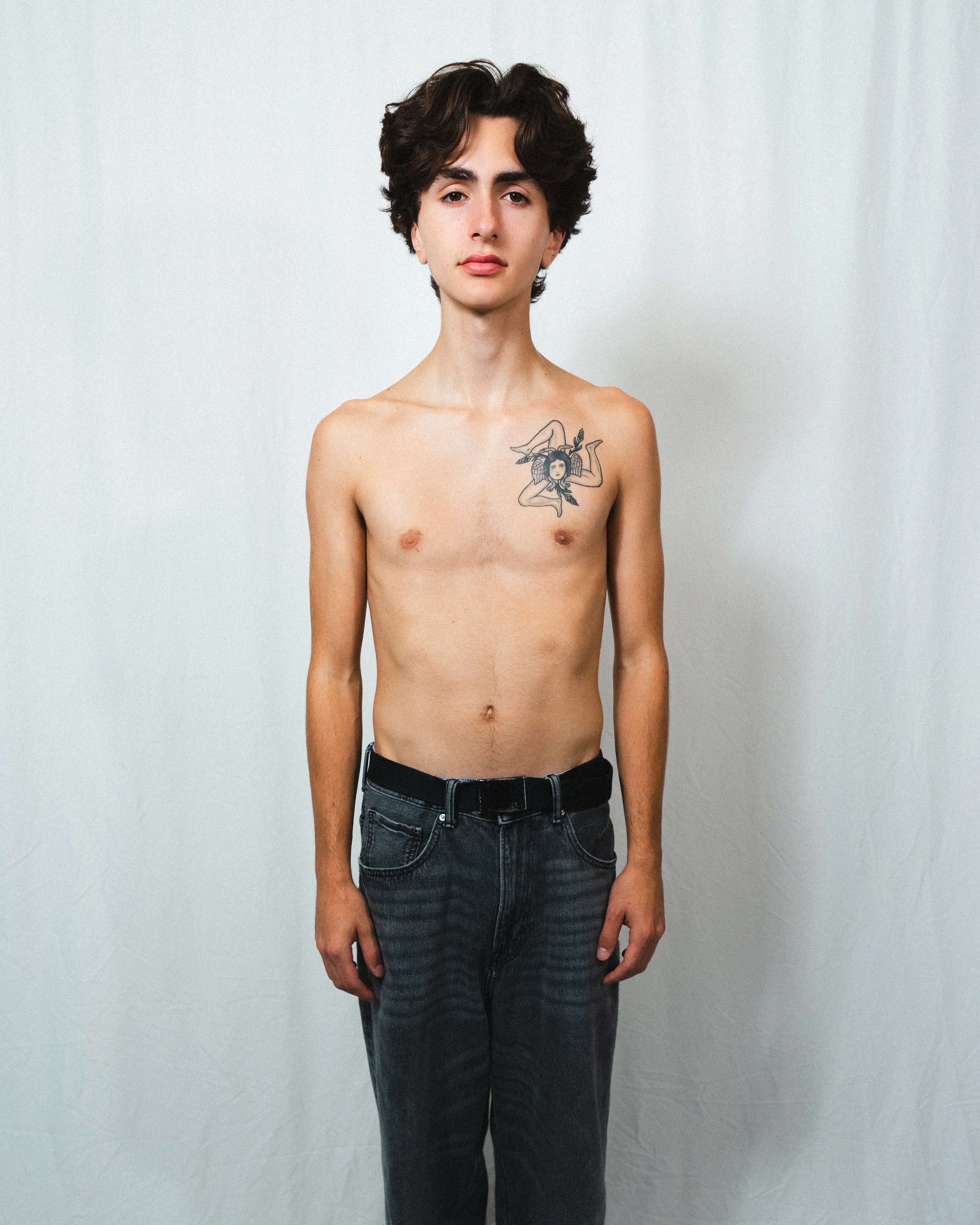 A young person with short, dark curly hair, standing shirtless against a plain white background, wearing black jeans with a belt, with a tattoo of a woman's face surrounded by foliage on the left side of the chest.