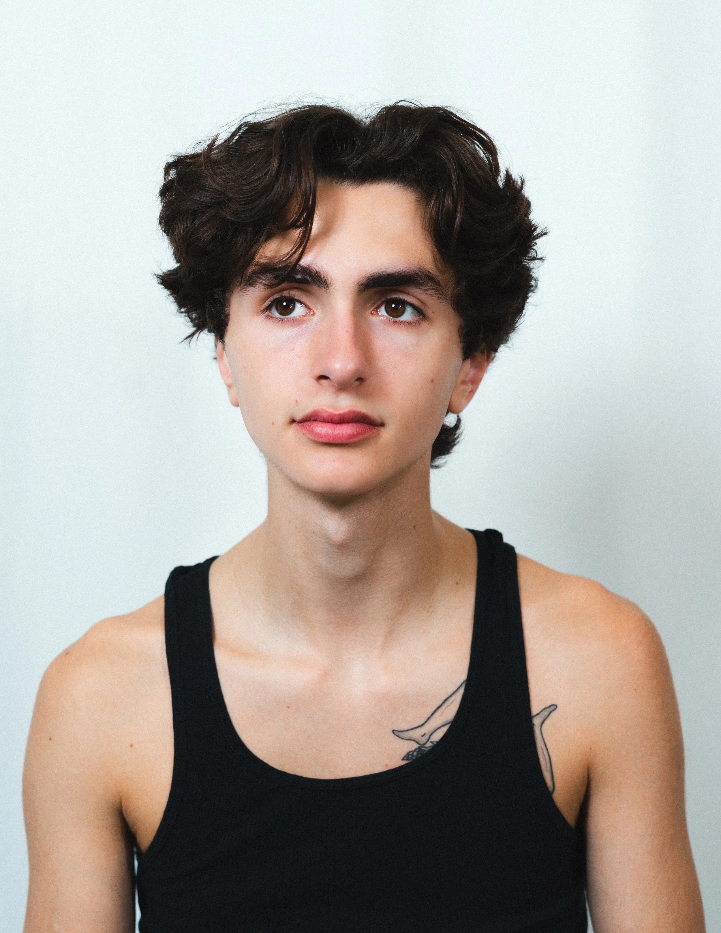 Portrait of a young man with short curly dark hair wearing a black tank top, looking slightly to his right, standing against a plain light background.