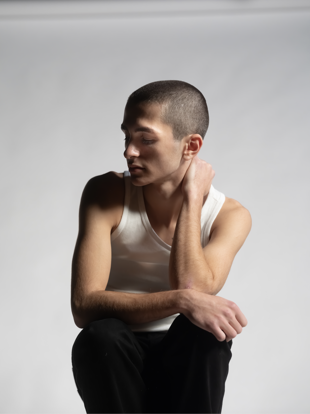 Young man wearing a white tank top and black pants sitting in a contemplative pose with his hand on his neck, against a plain light gray background.