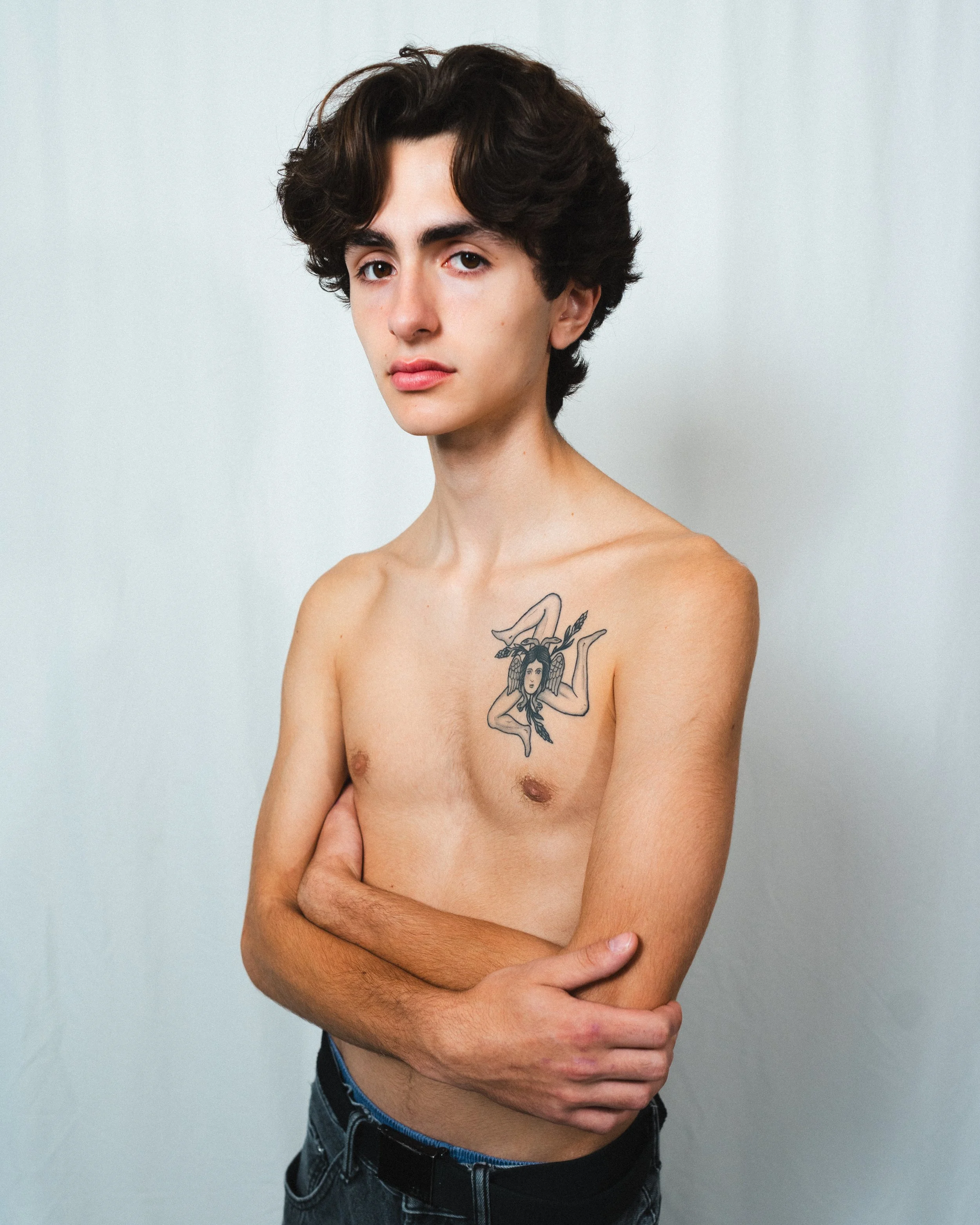 A young man with dark wavy hair and light skin, shirtless, with a tattoo of a woman with long hair and feather accessories on his left chest, standing against a plain light background.