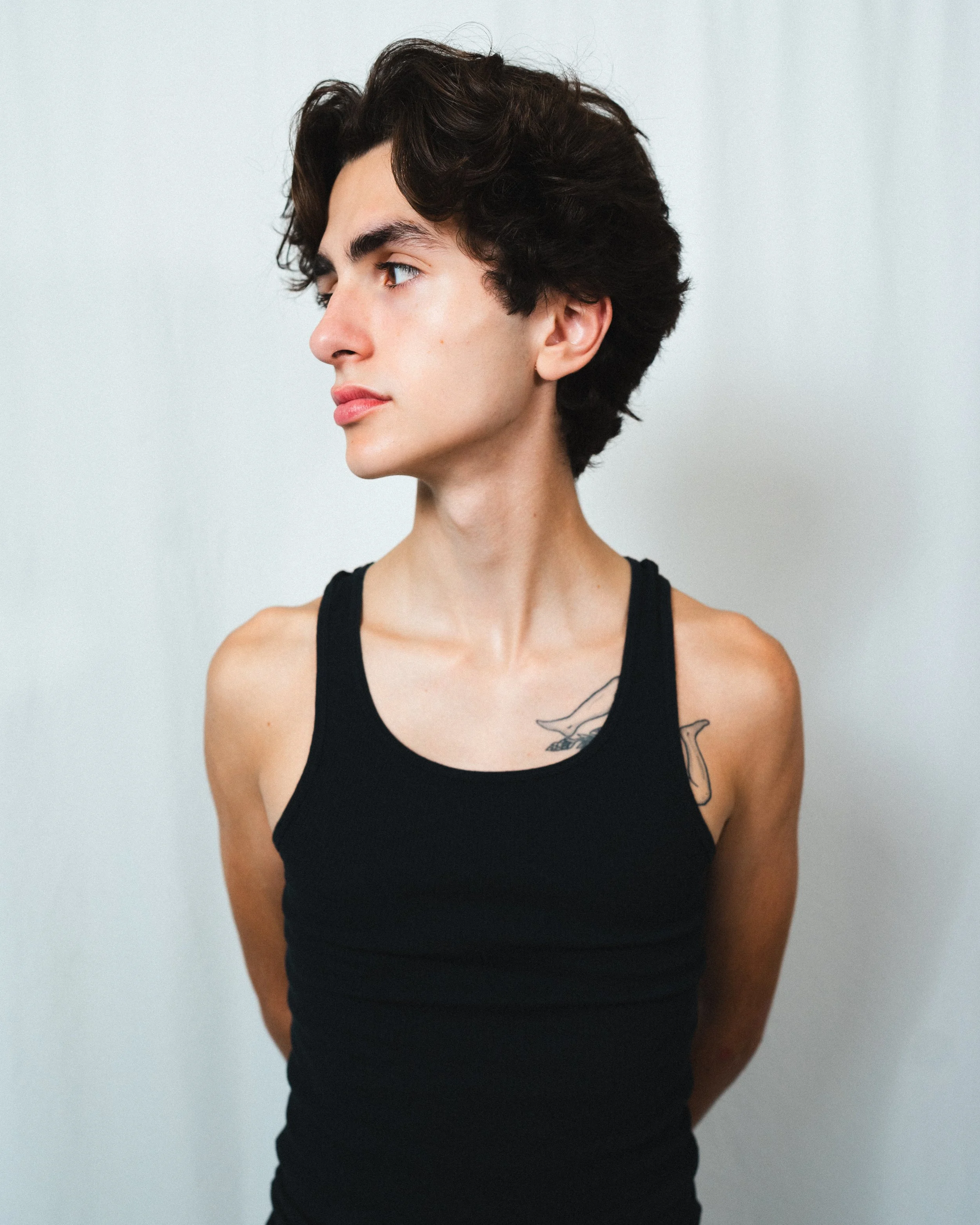 Portrait of a young person with short dark hair wearing a black tank top, with tattoos visible on their shoulder and neck, standing against a plain light background.