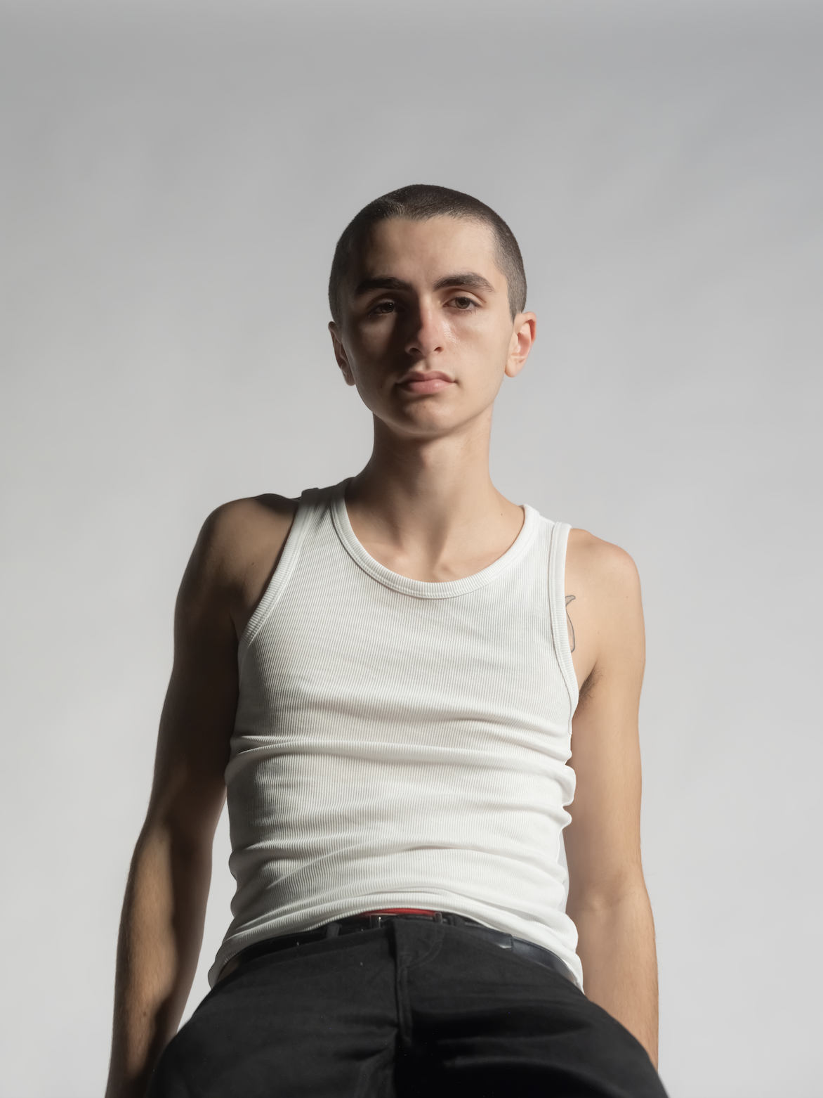 A young man with a shaved head wearing a white tank top and black pants, standing against a plain gray background.