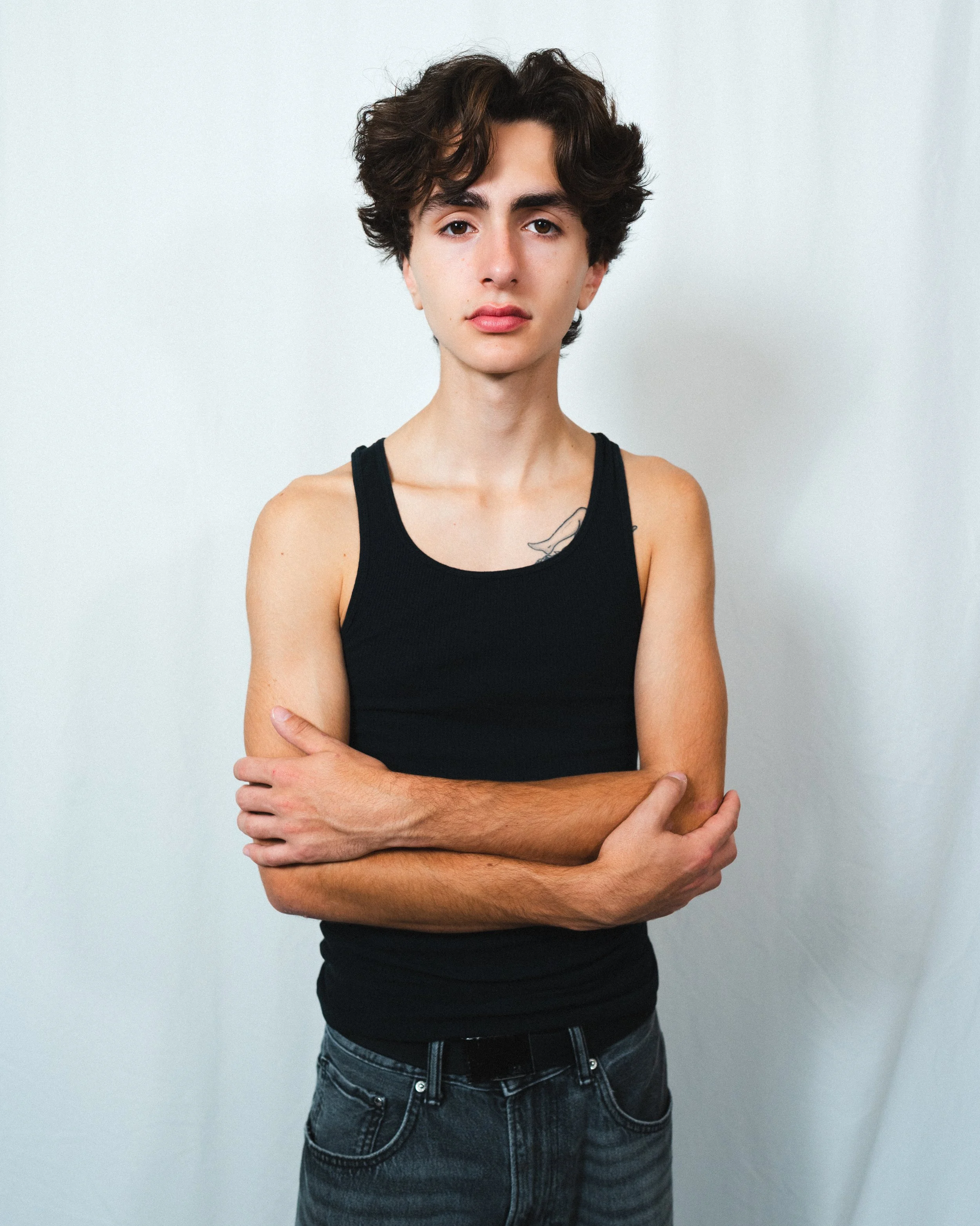 Young person with short curly hair, wearing a black tank top and ripped jeans, standing against a plain white background with arms crossed.