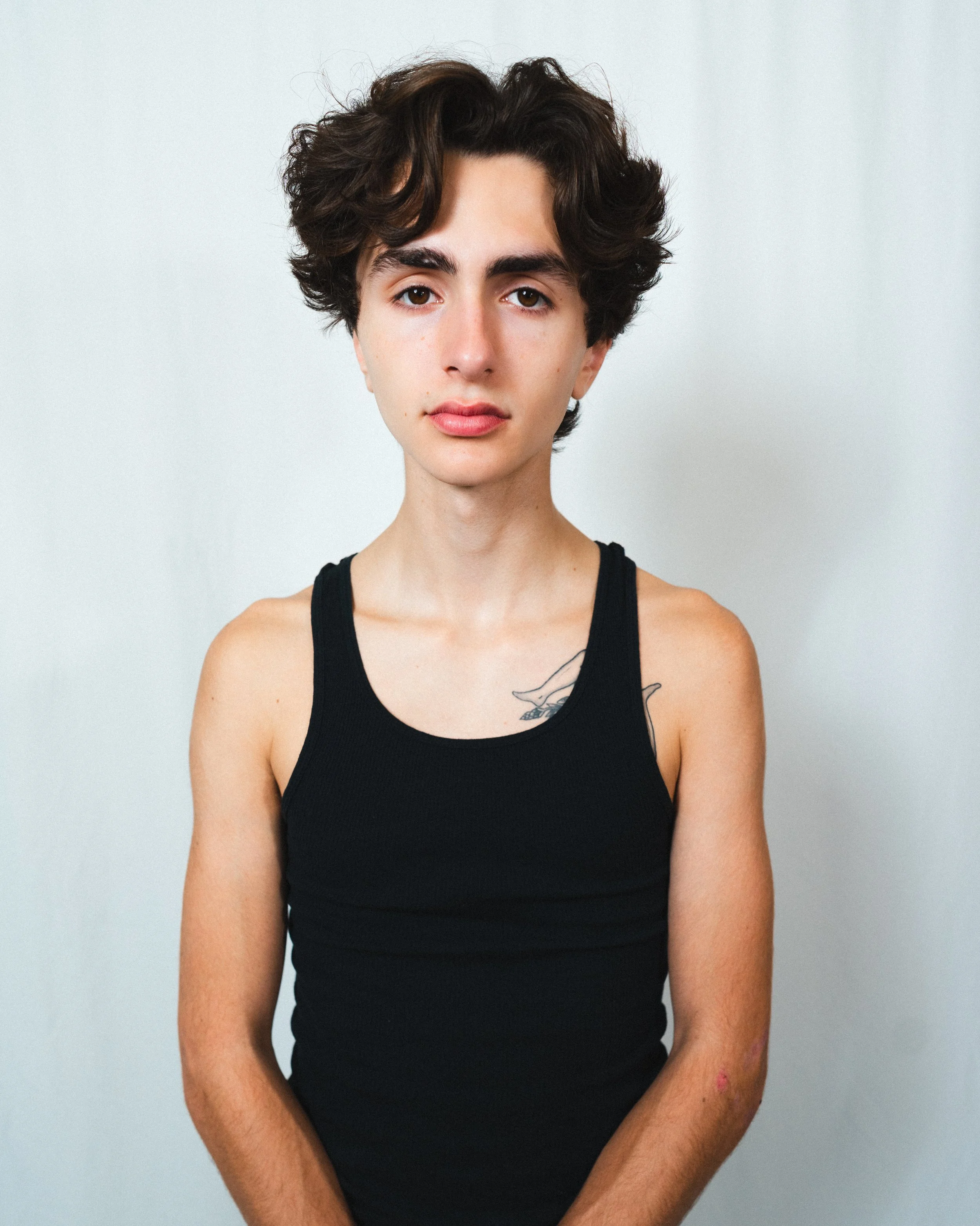 Portrait of a young person with short, wavy dark hair, wearing a black sleeveless top, standing against a neutral background.