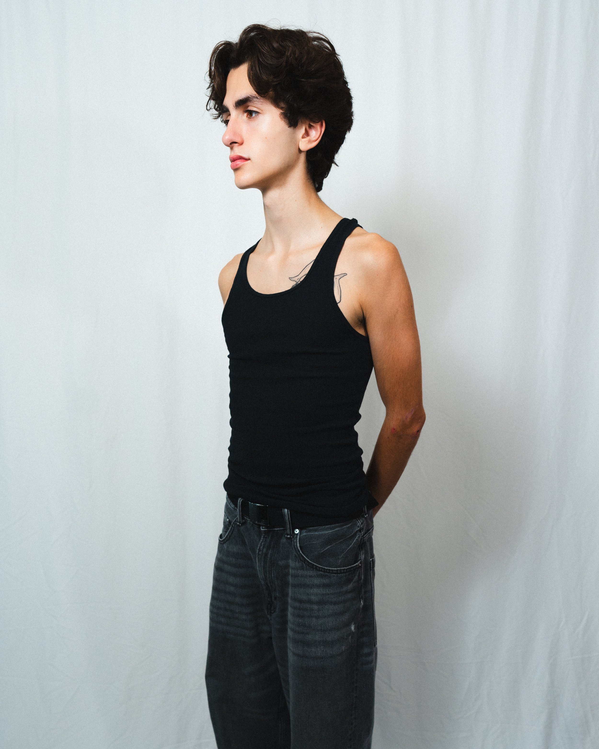 A young person with short dark hair, wearing a black tank top and black jeans, standing with arms behind their back against a plain white background.