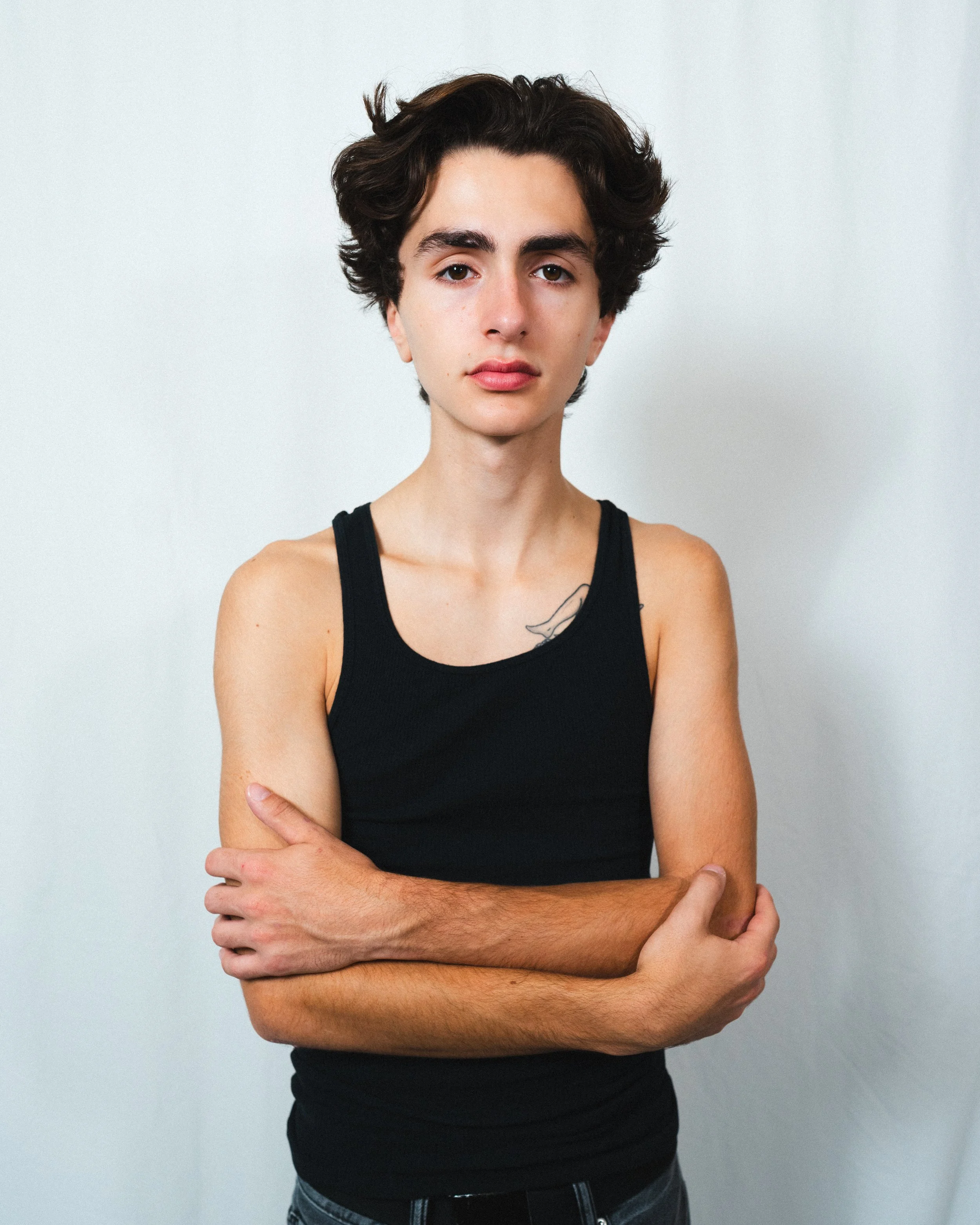 Portrait of a young man with short curly dark hair, wearing a black tank top, standing against a plain light background with arms crossed.