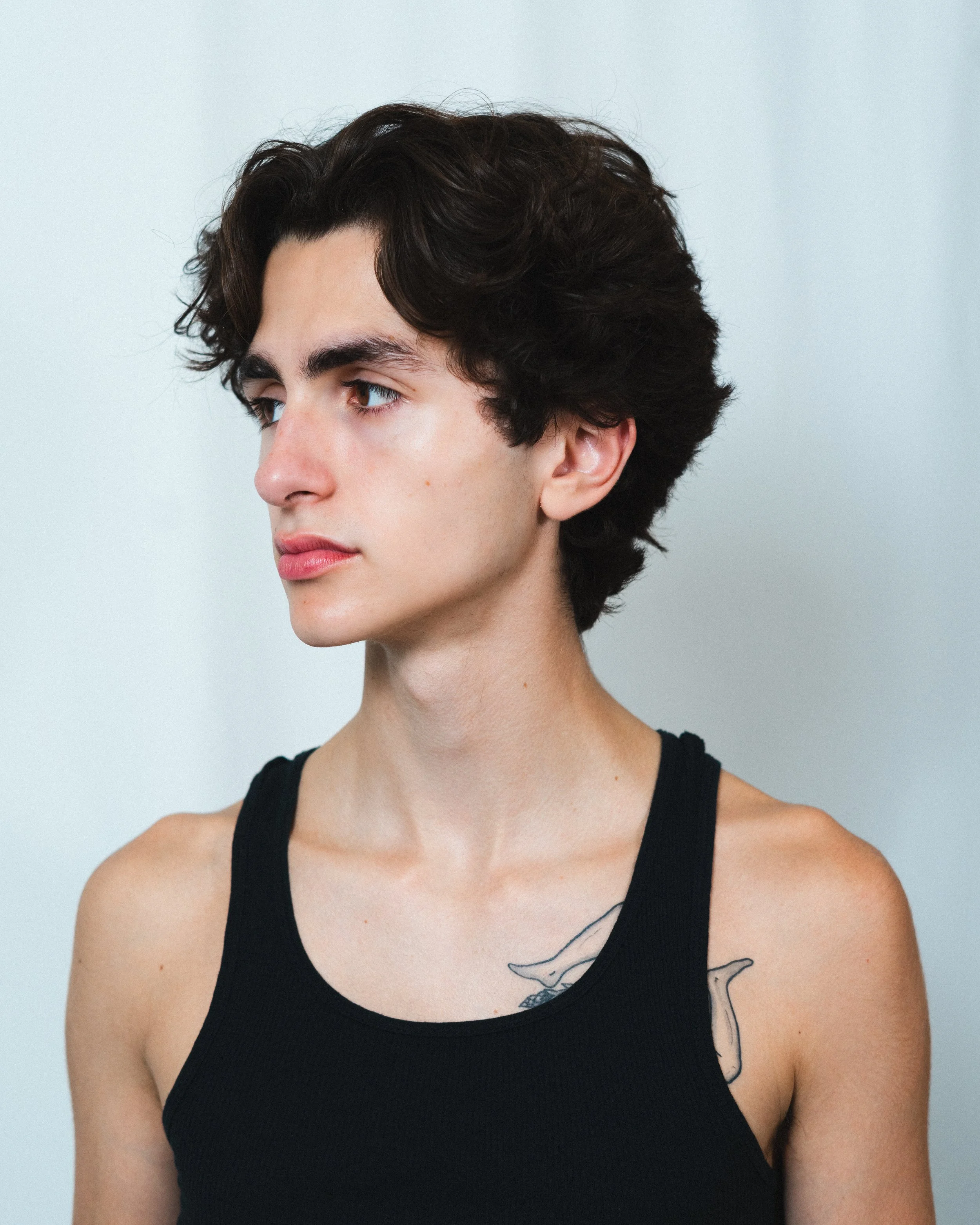 A young man with short, dark wavy hair and light skin, wearing a black sleeveless top, is looking to his left against a plain light background.