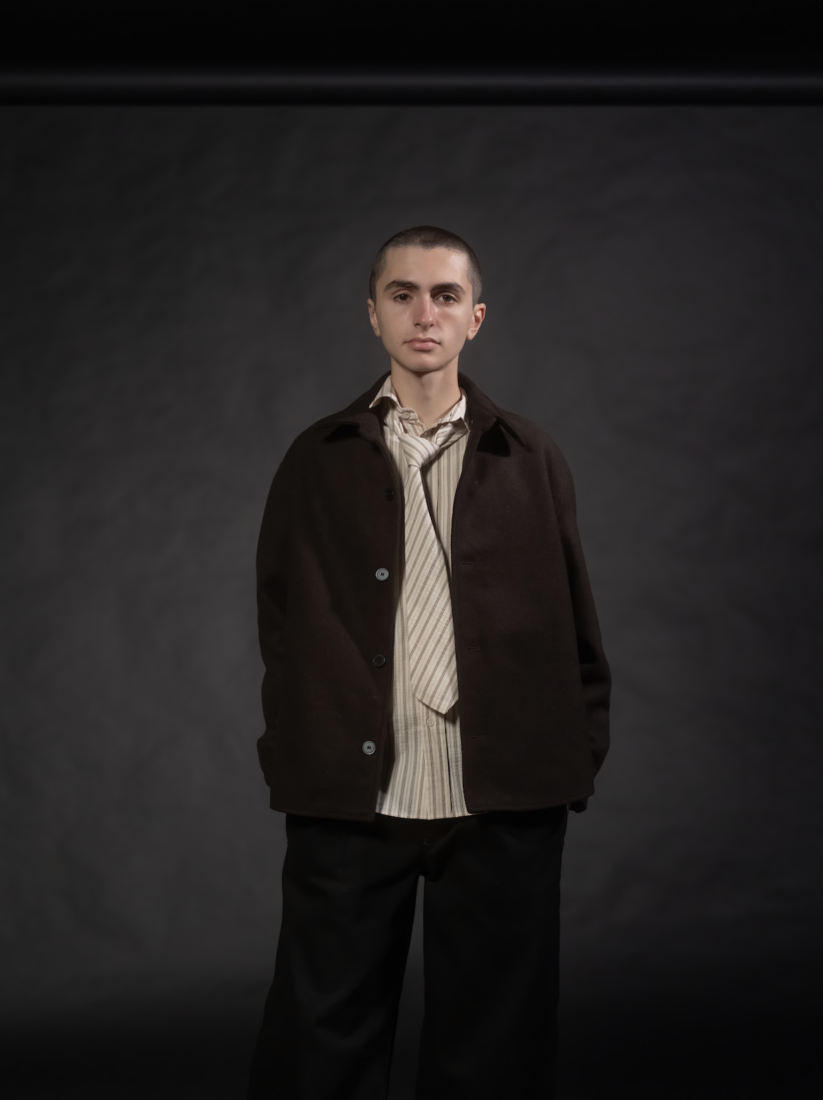 A young man with short hair, wearing a dark coat over a striped shirt and a loosened tie, standing against a plain dark background.