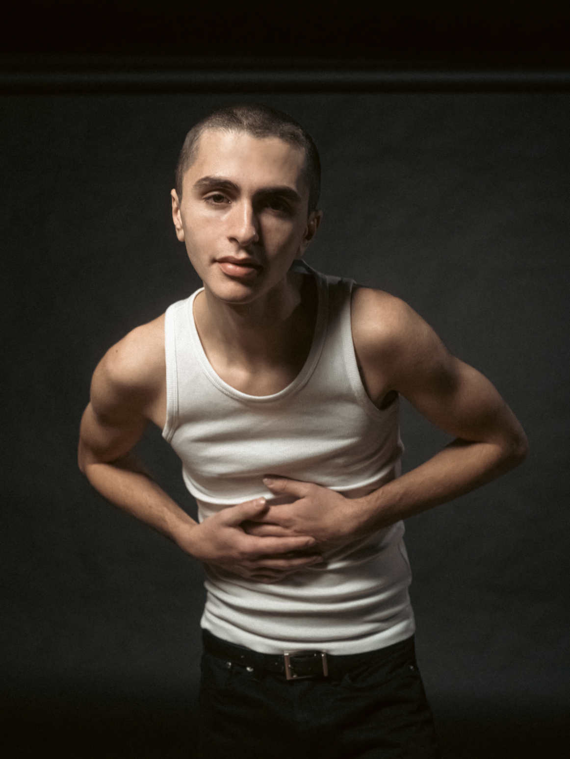 Person with short hair and white tank top holding stomach, looking at camera against dark background.